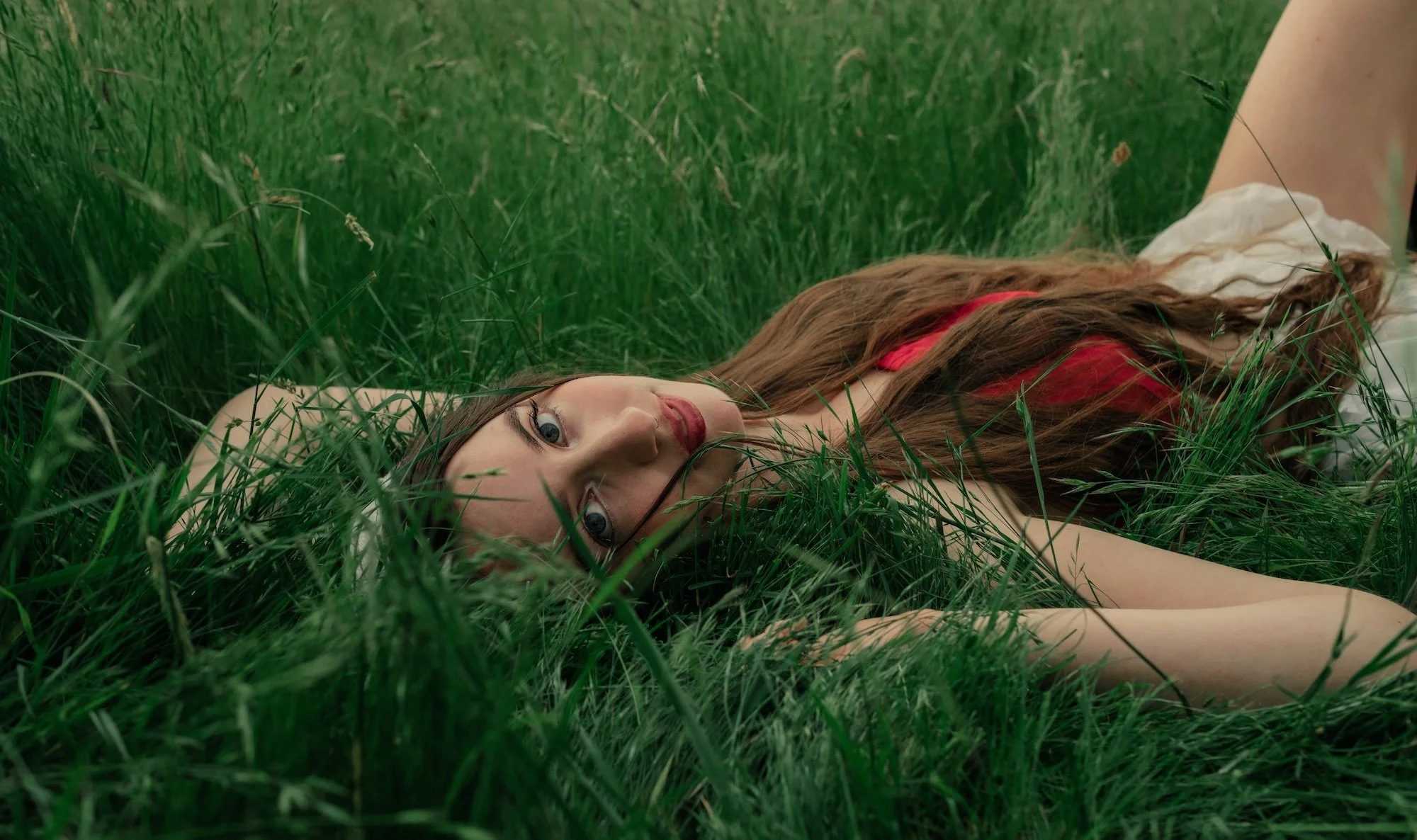 A woman lies in tall green grass, gazing toward the camera with a calm expression, partially surrounded by lush foliage