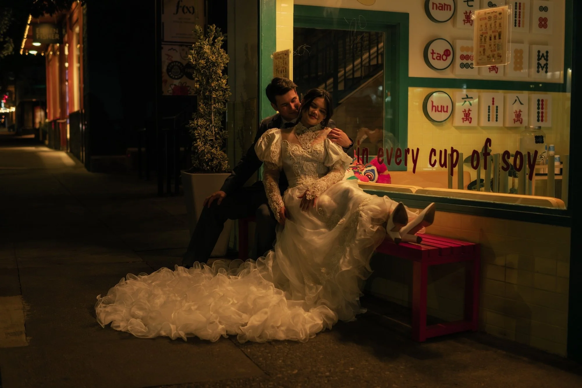 Bride and groom sitting together outside at night, illuminated by warm street and storefront lights