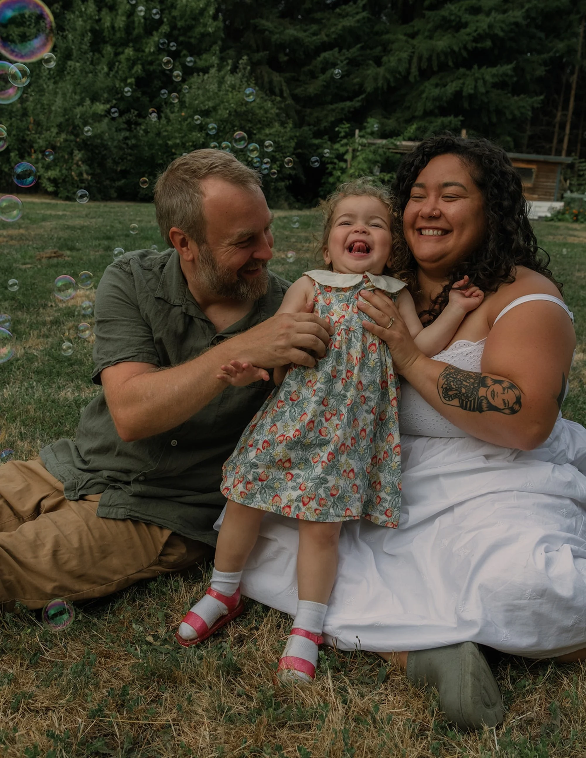 A family of three sits on grass outdoors, smiling and playing together as their young child laughs with bubbles floating around them