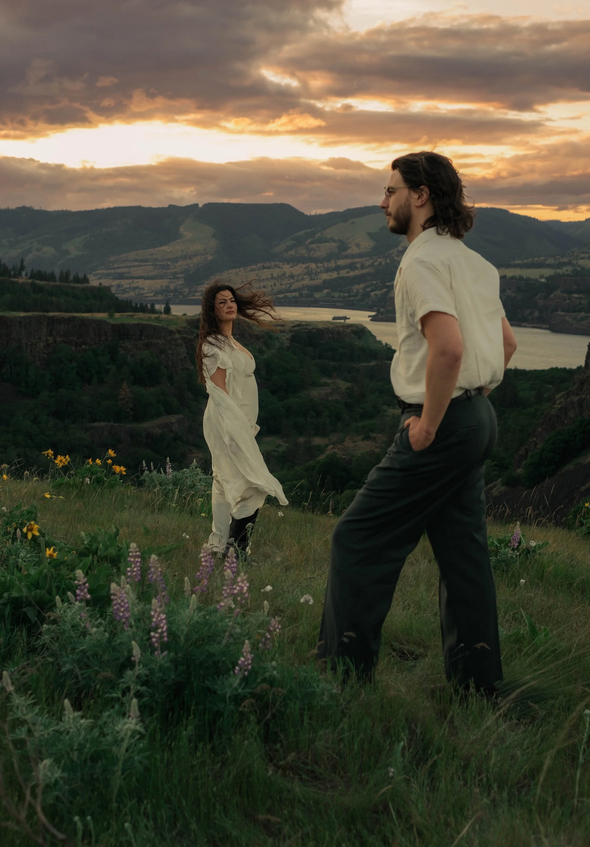 Couple on a grassy hillside at sunset, with a woman in a flowing white dress and a man in the foreground overlooking a river valley