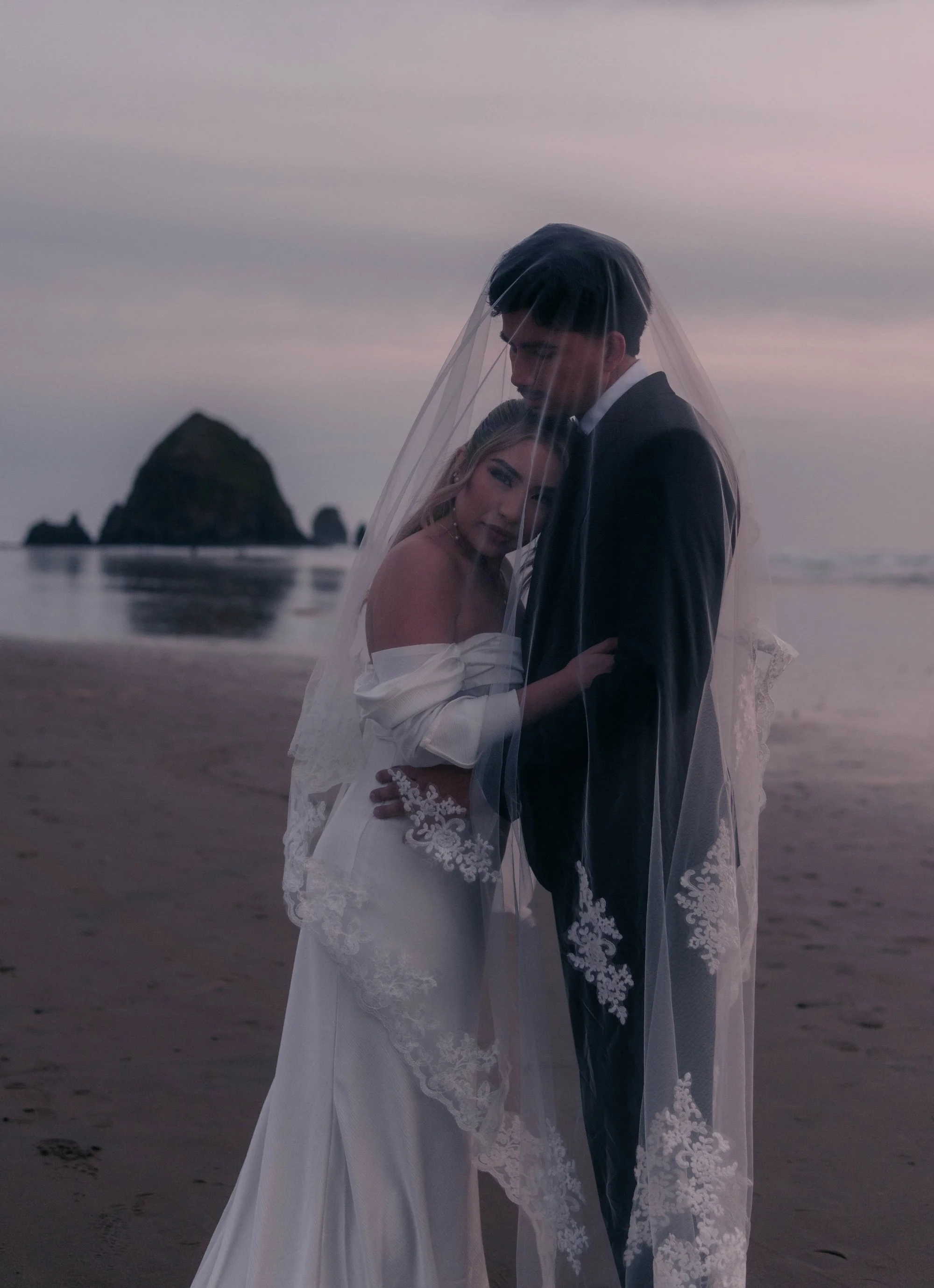 A bride and groom embrace on a misty beach, partially covered by a sheer veil, with a large sea stack visible in the background