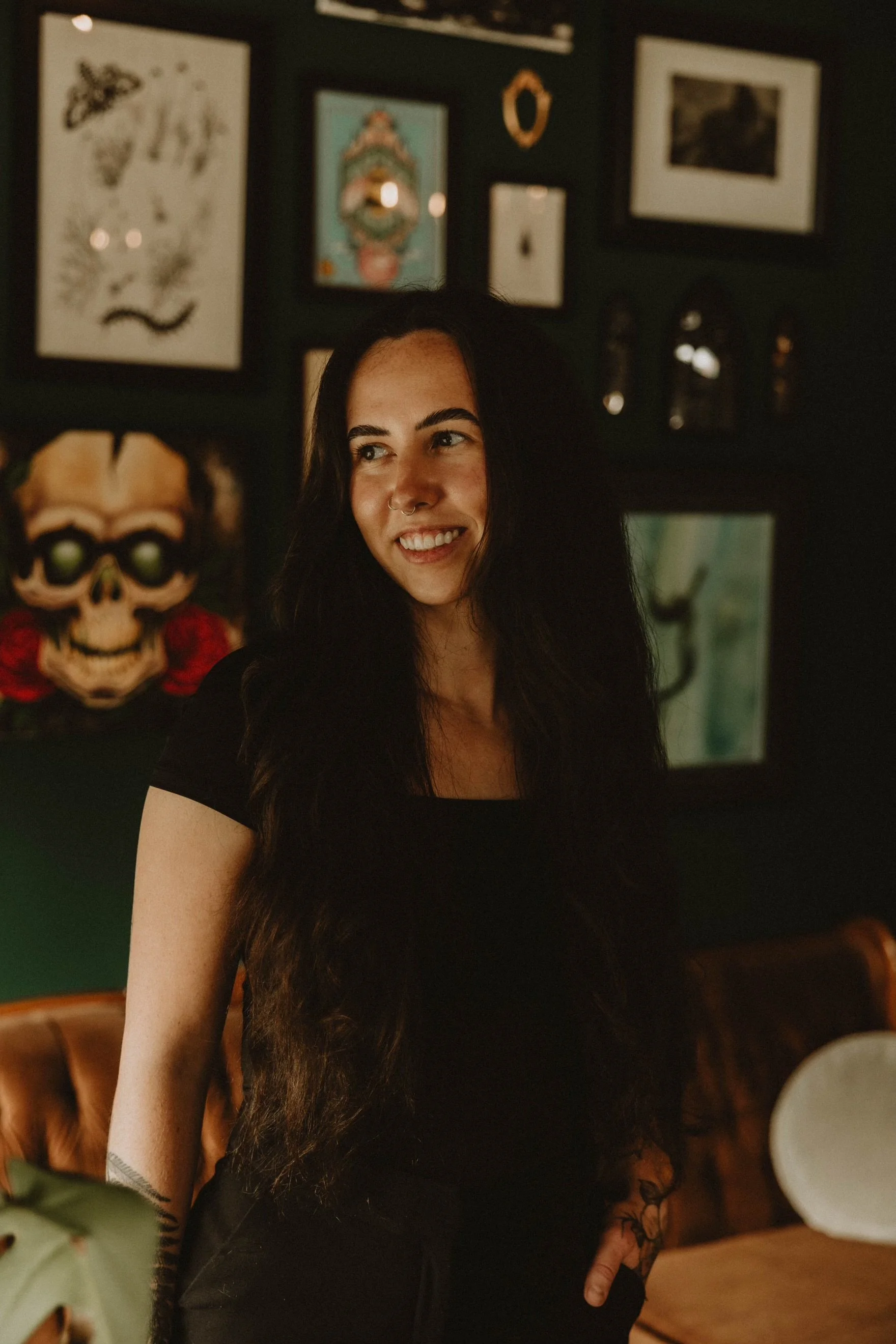 Portrait of a woman with long dark hair, smiling, standing in a dimly lit room with artwork on the dark wall behind her, including a skull, framed pictures, and decorative objects.