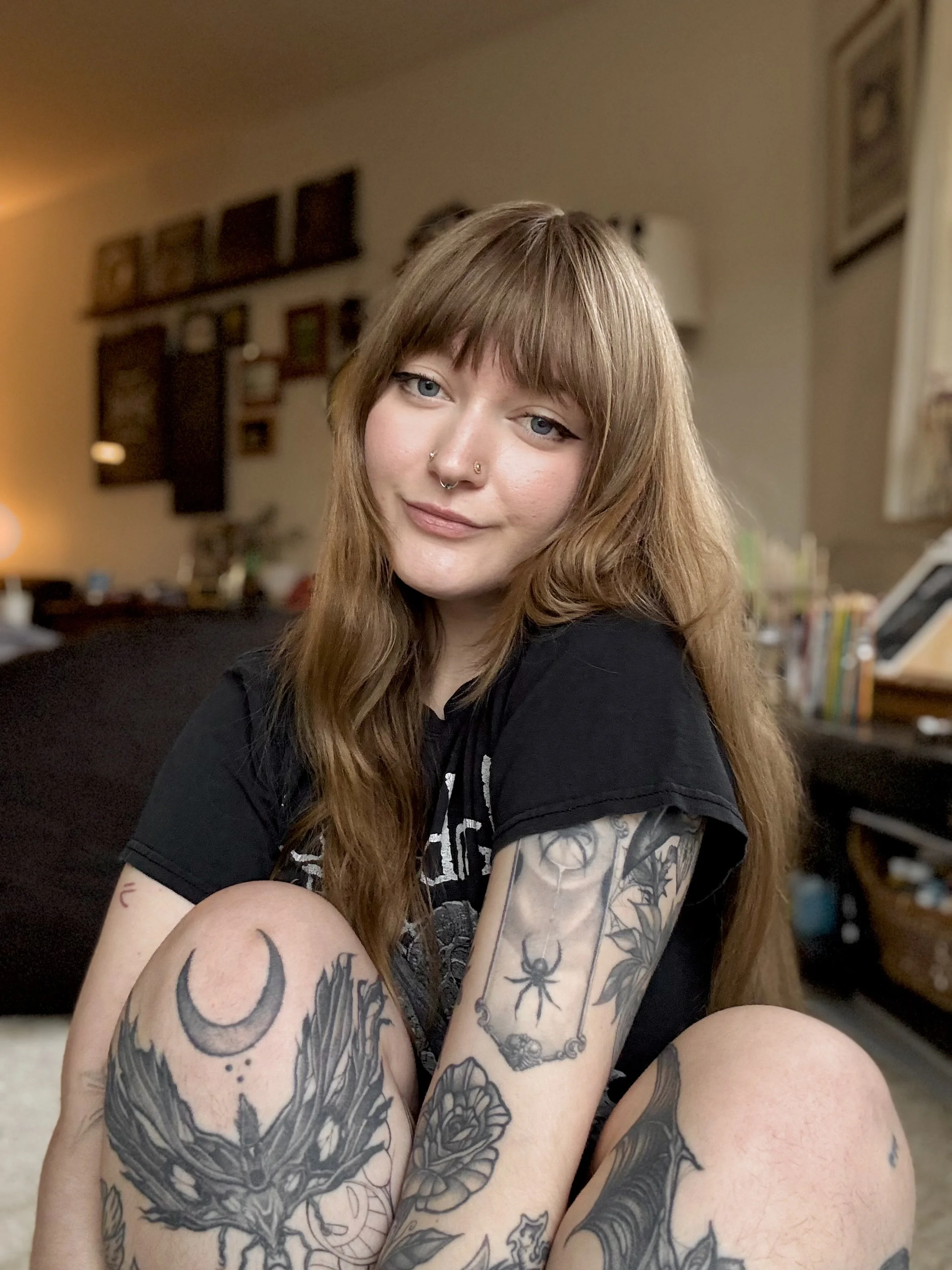 A young woman with long, wavy reddish-brown hair and tattoos on her arms, sitting indoors with a neutral expression, wearing a black shirt.