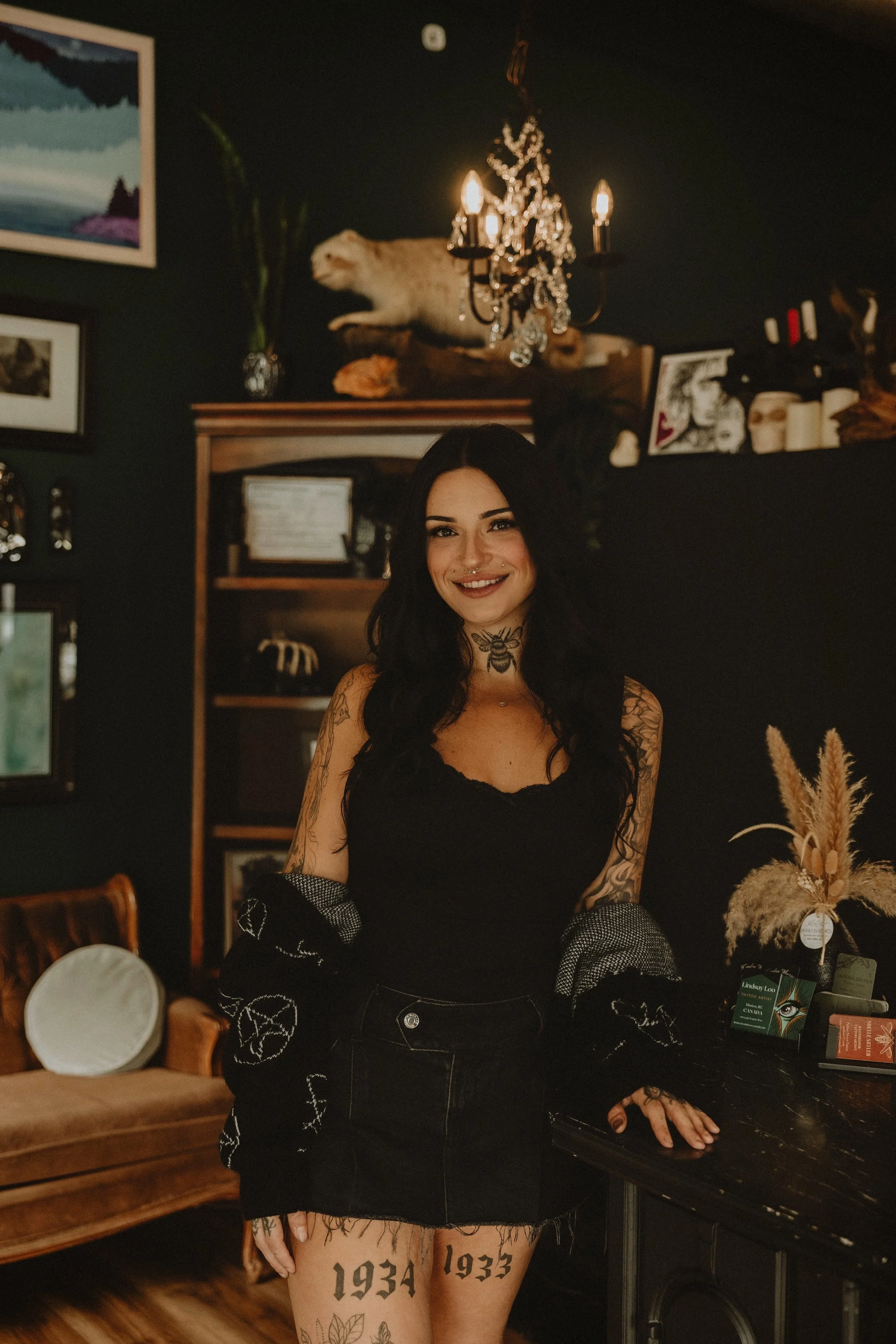 A woman with long black hair, tattoos, and a smile, standing in a dimly lit room with dark walls, framed photos, a chandelier, a shelf with decorative items, and a plush bench. There are decorative plants and artwork in the background.