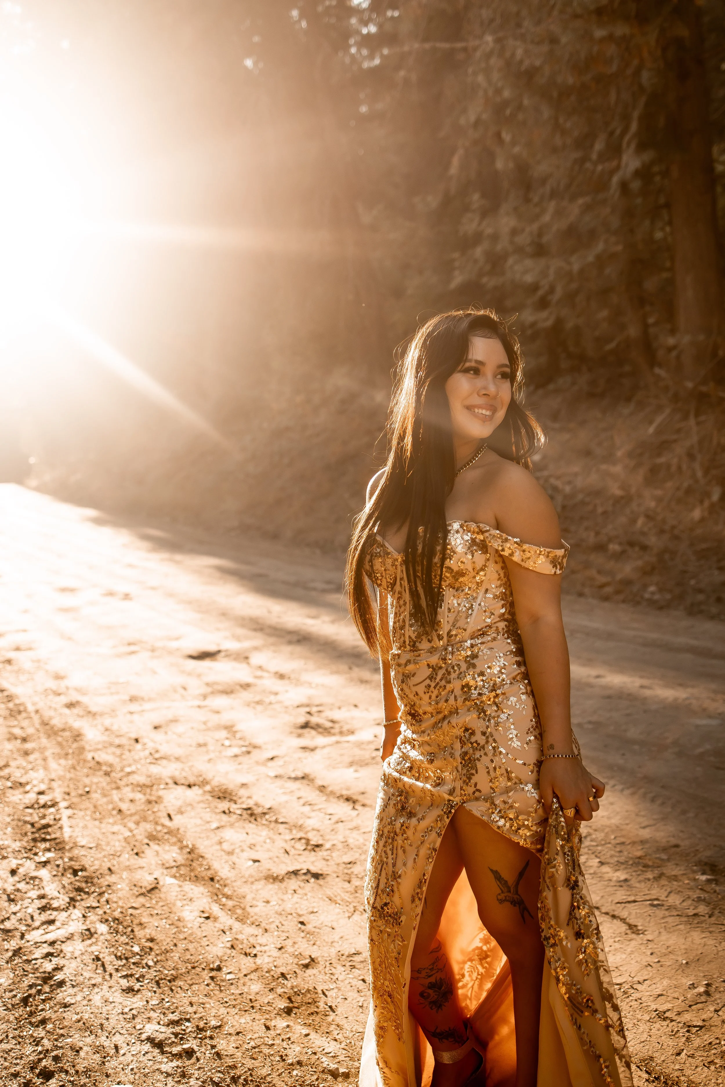 A woman with long brown hair wearing a gold, embellished gown with a slit, standing on a dirt path with a rocky hillside in the background during golden hour sunlight.