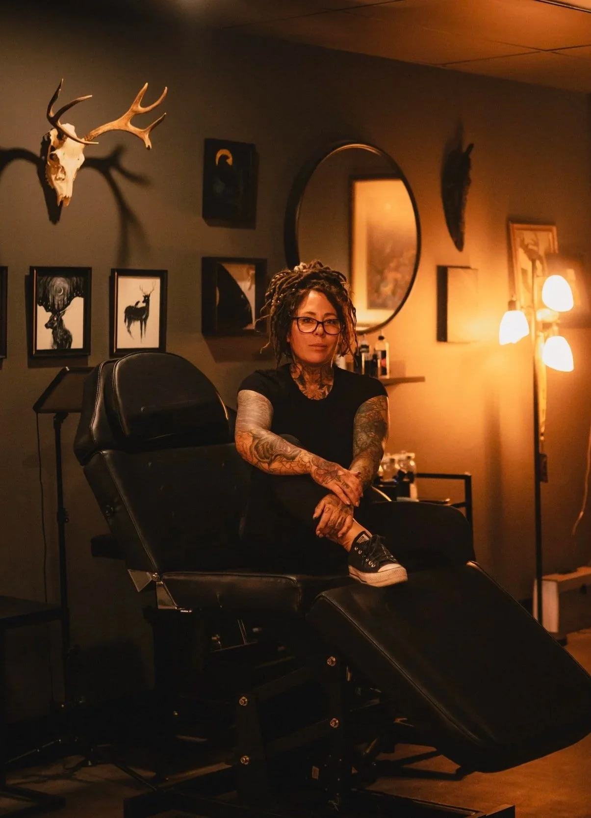 A woman with dreadlocks, glasses, and tattoos sitting on a black salon chair in a tattoo studio or barber shop with framed pictures, animal skull, and wall art behind her, illuminated by warm lighting.