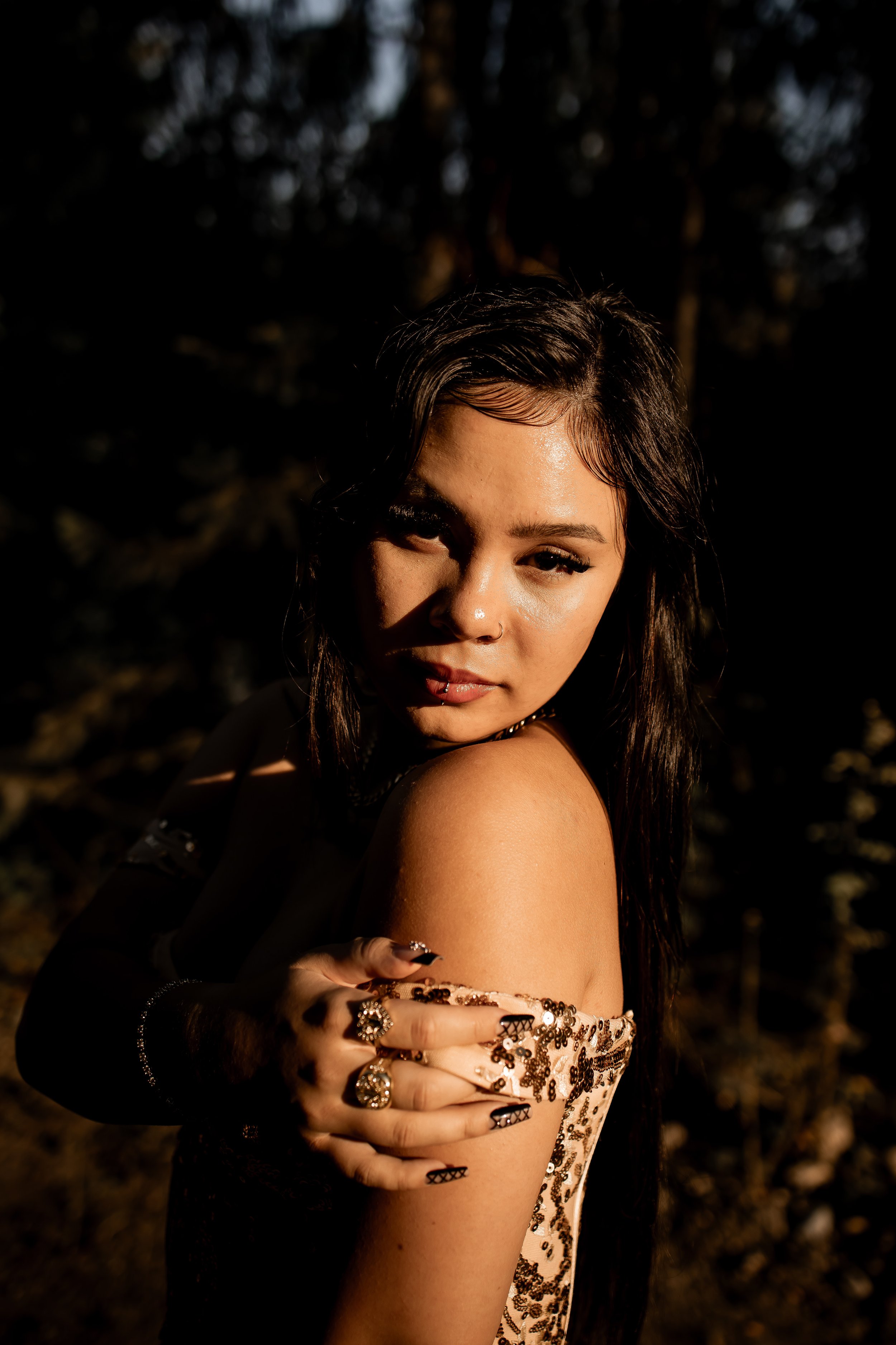 A young woman with wet dark hair and tattoos on her arm, wearing a floral off-shoulder top, is posing outdoors at sunset or sunrise with a dark background and trees.