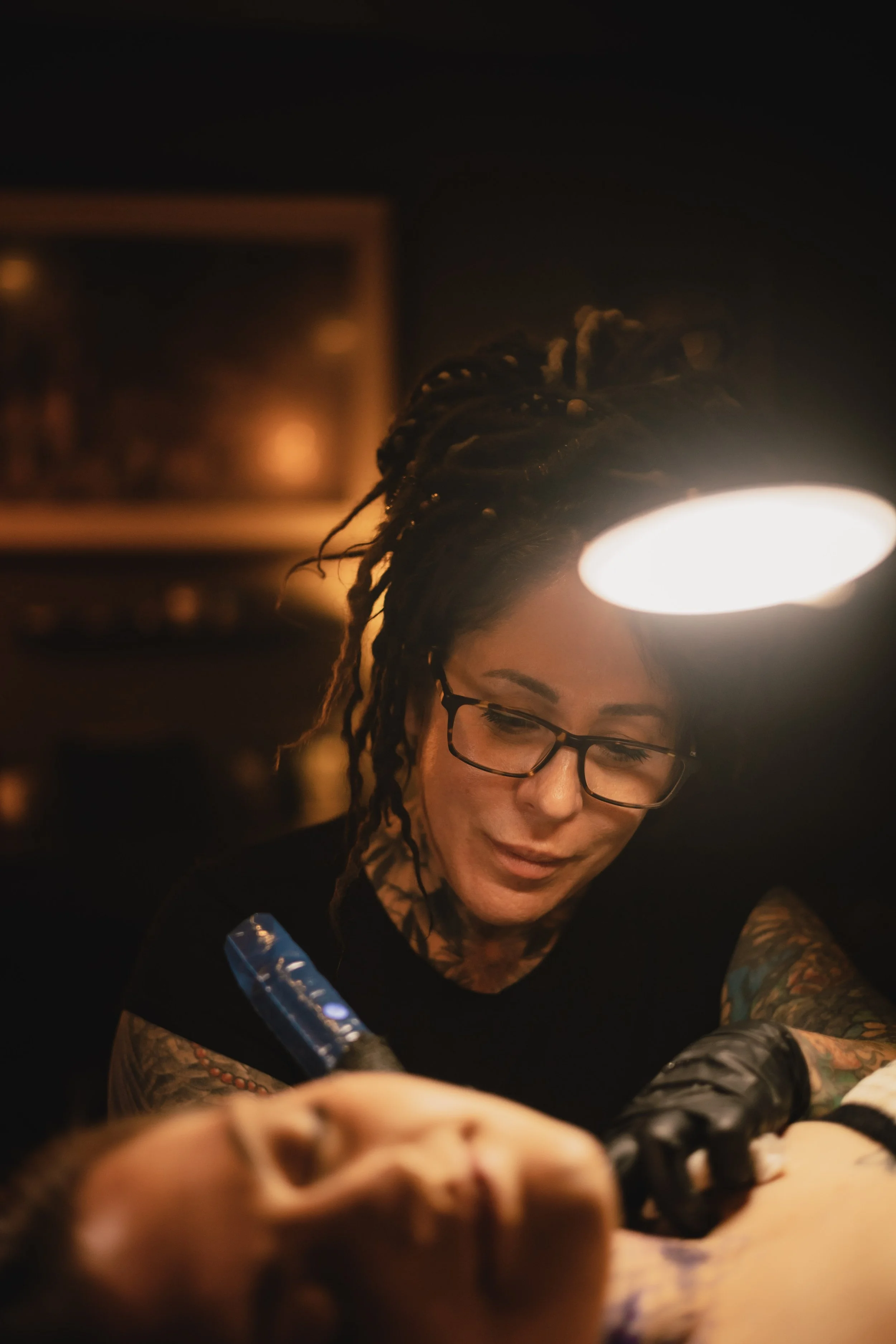 A woman with glasses and tattoos on her arms is lighting a tattoo needle in a dimly lit room.