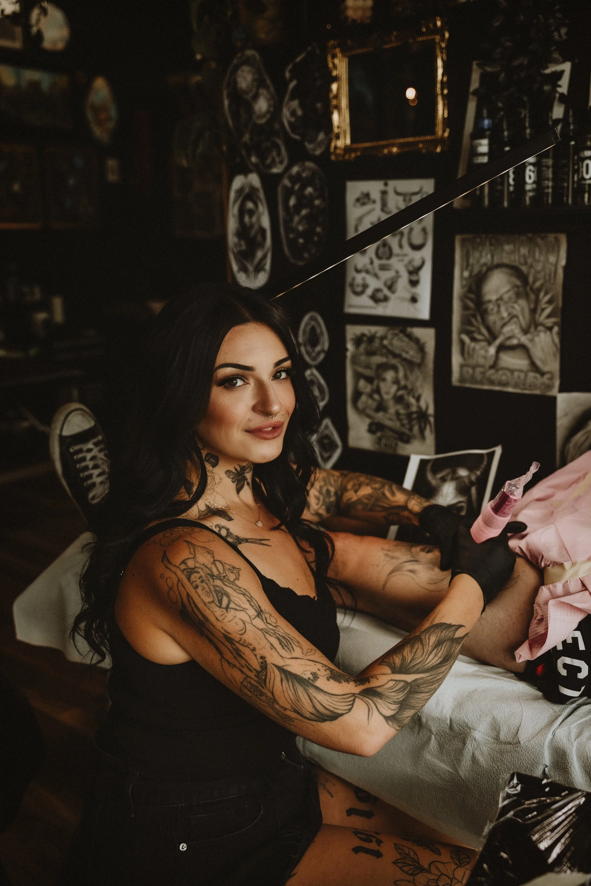 A woman with black hair, tattoos on her arms and shoulder, sitting on a tattoo table, smiling, with tattoo artwork visible on her skin, and a tattoo artist in the background.