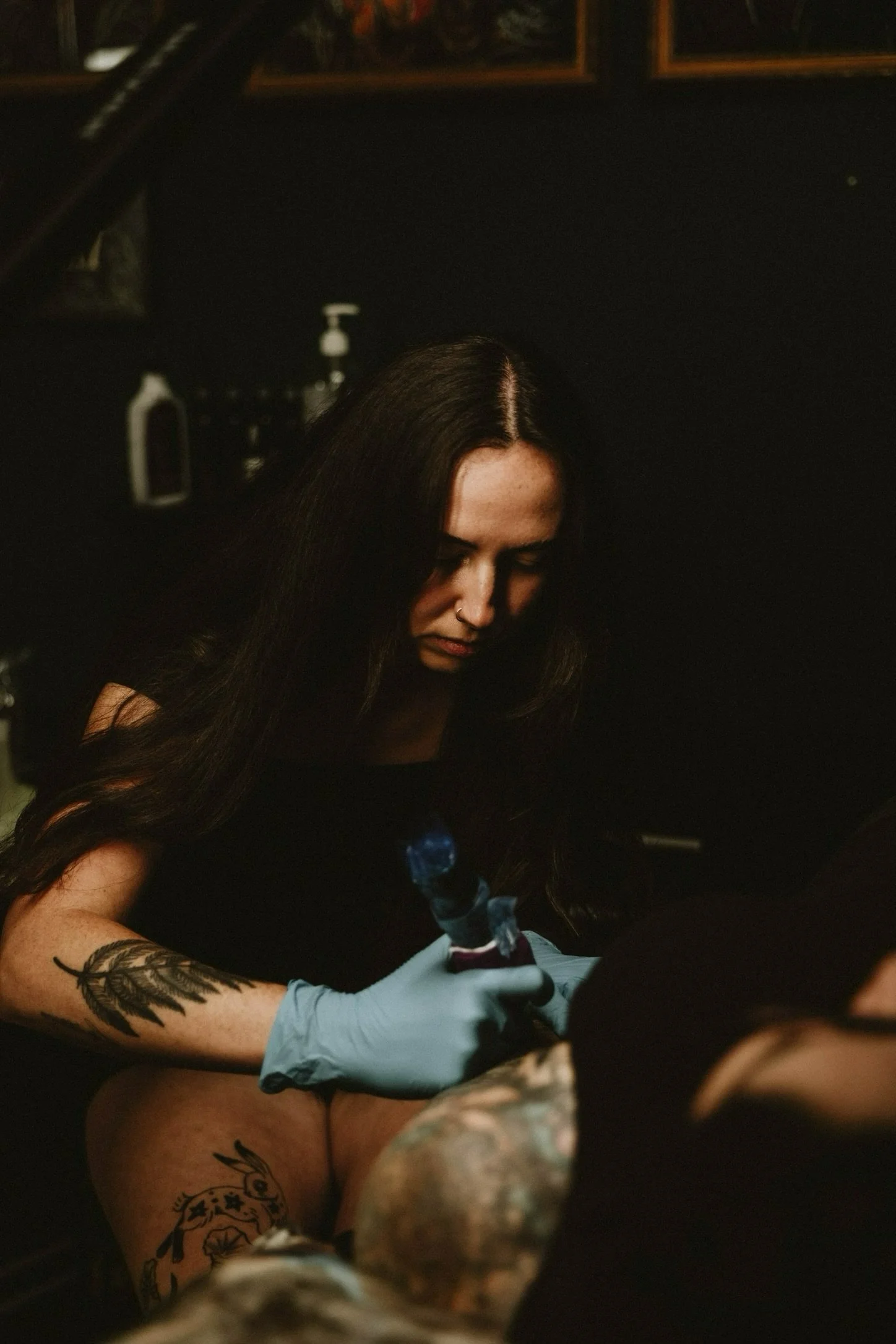 A woman with long dark hair getting a tattoo by a tattoo artist. The woman has tattoos on her right arm and thigh and is wearing gloves while the artist holds a tattoo gun. The setting is a tattoo studio with a dark background.