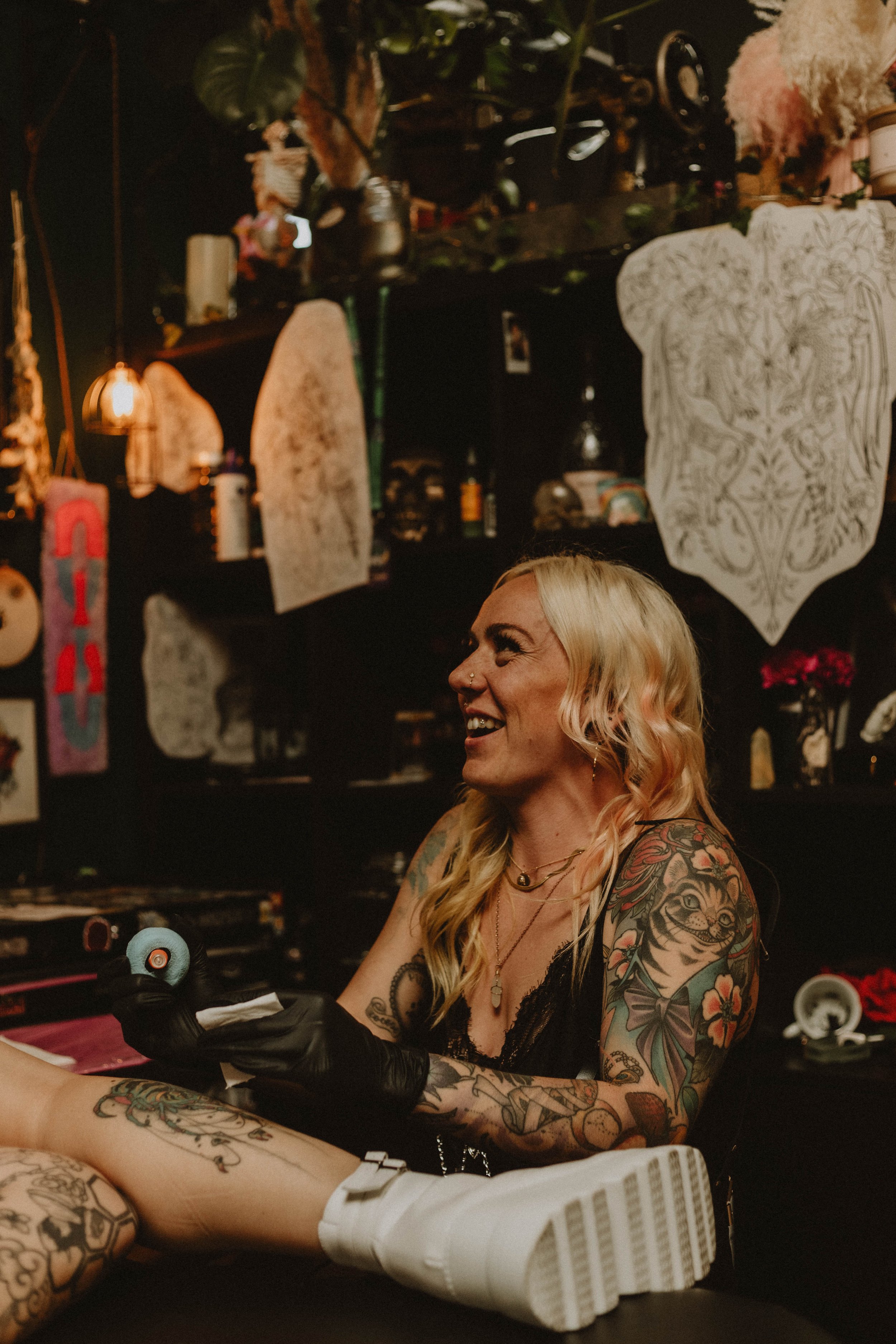 A woman with long blonde hair and tattoos on her arms and shoulder, smiling while inking a client's arm in a tattoo parlor. The woman is wearing black gloves and a tank top, with various colorful tattoos visible on her arms and shoulder. The background shows shelves with various decorations, plants, and tattoo-related items.