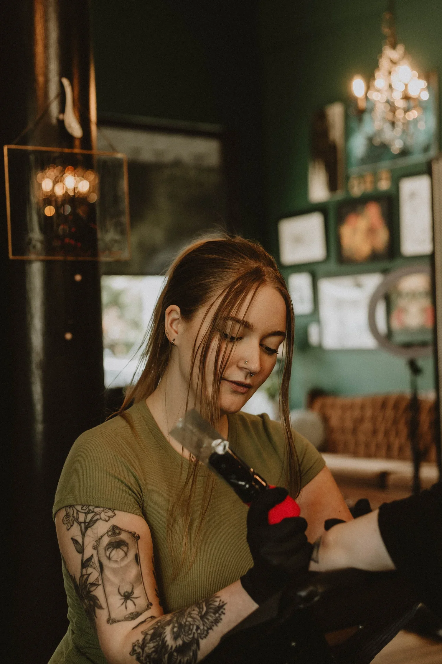 A woman with tattoos on her arms, wearing a green shirt, holds tools and is getting a tattoo on her arm in a tattoo parlor.