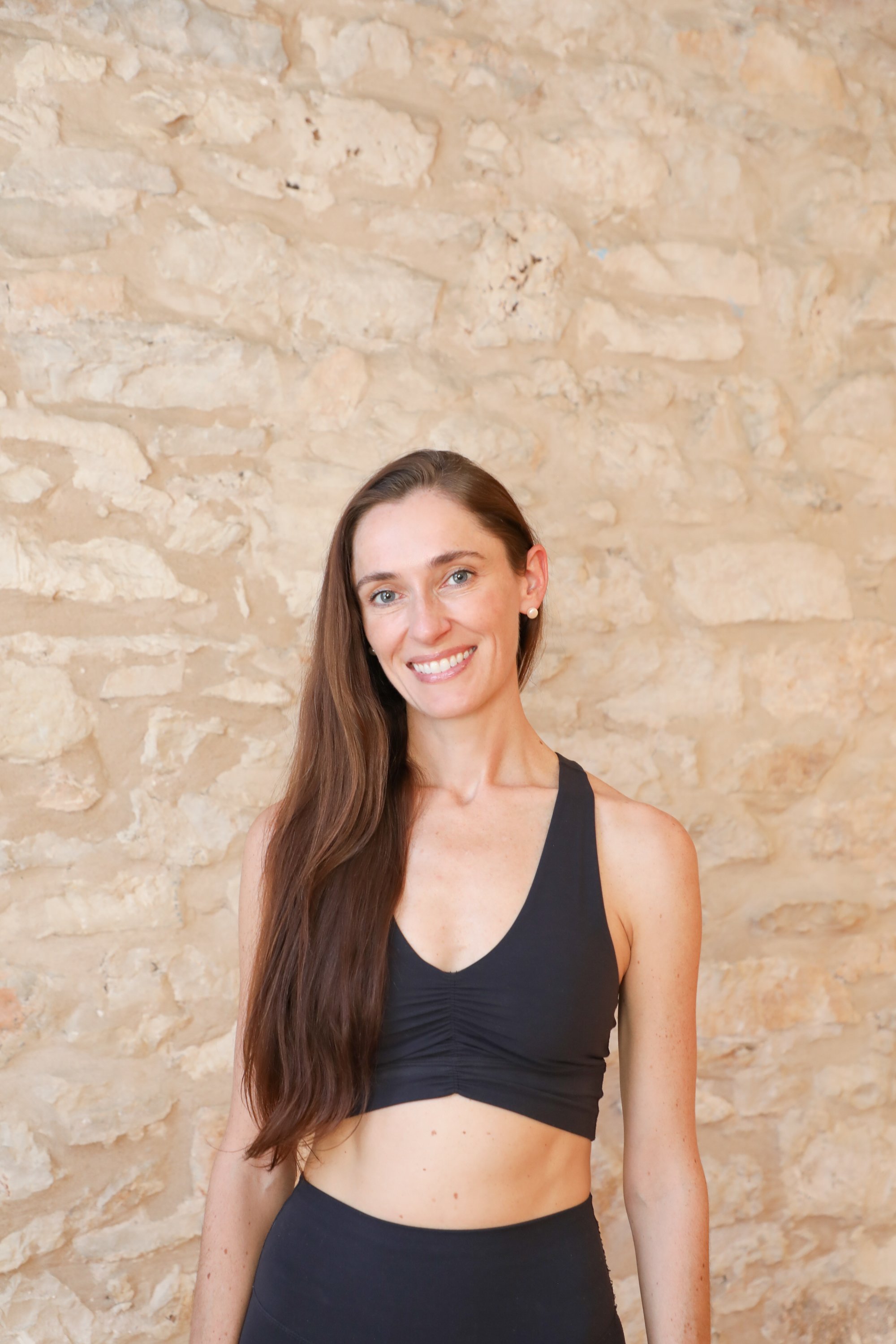 A woman with long brown hair wearing a black sports bra and black sports leggings standing in front of a beige stone wall, smiling at the camera.