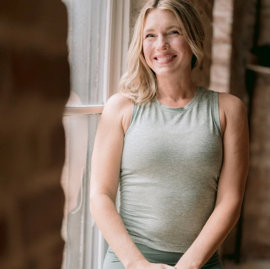 A woman with blonde hair smiles and looks out a window in a bright room, wearing a sleeveless gray top.