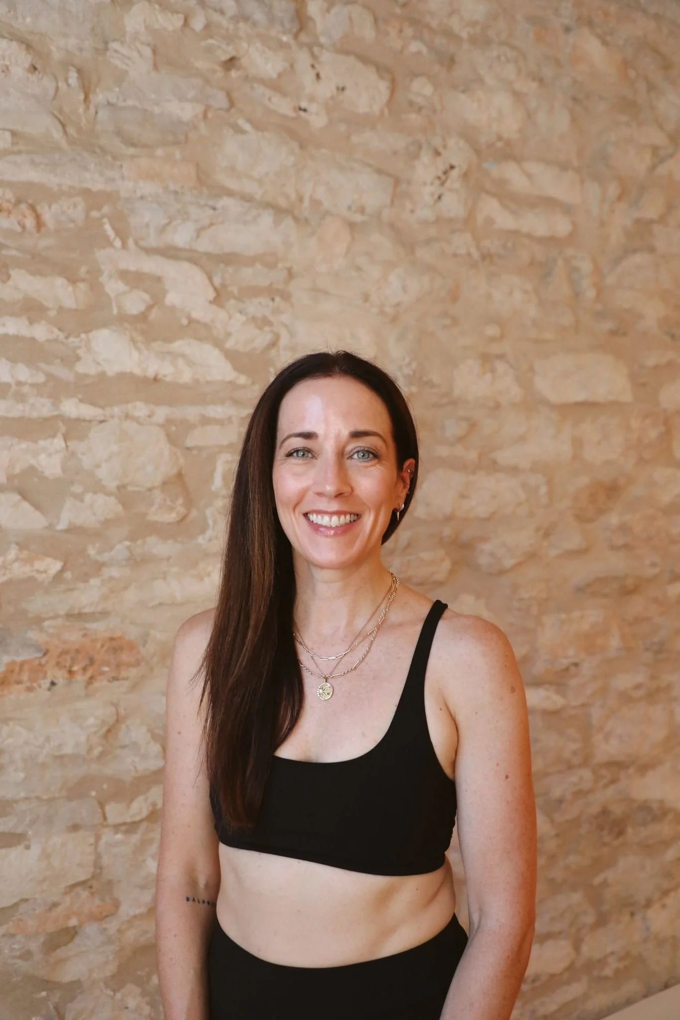 A woman with long dark hair and light skin smiling, wearing a black sports bra and layered necklaces, standing in front of a beige stone wall.