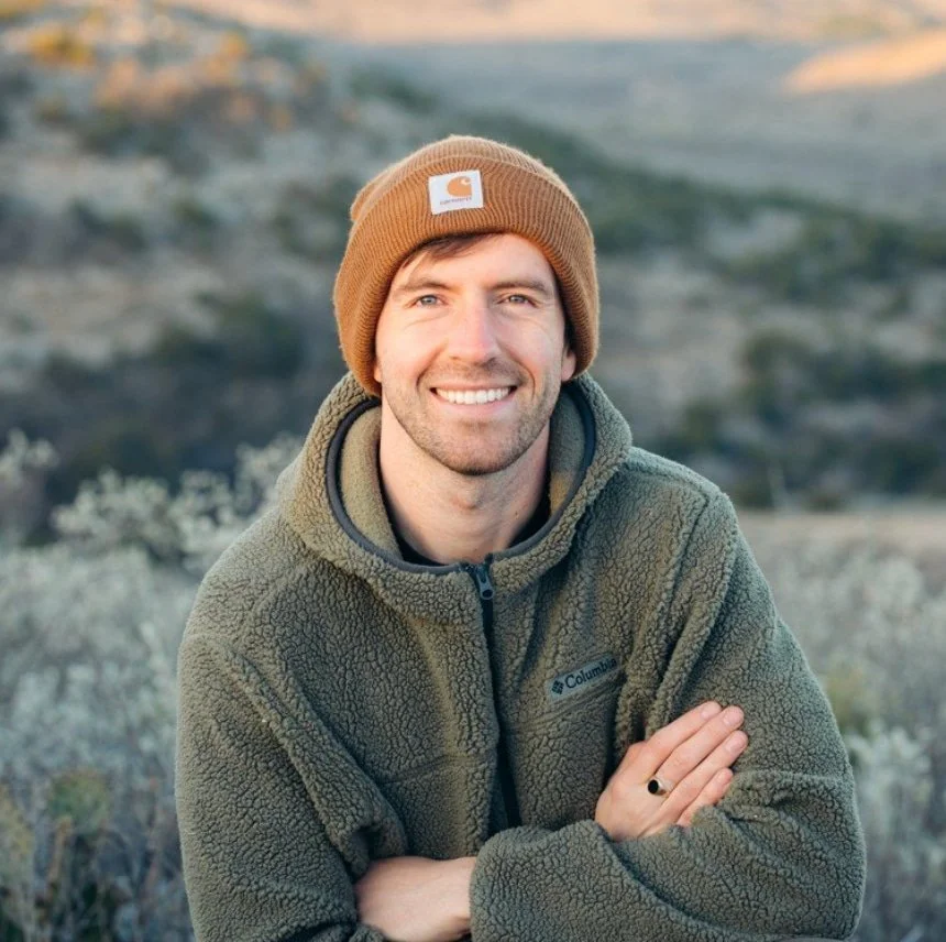 A smiling man with light skin, wearing a brown beanie and a green fleece jacket, standing outdoors in a natural landscape.