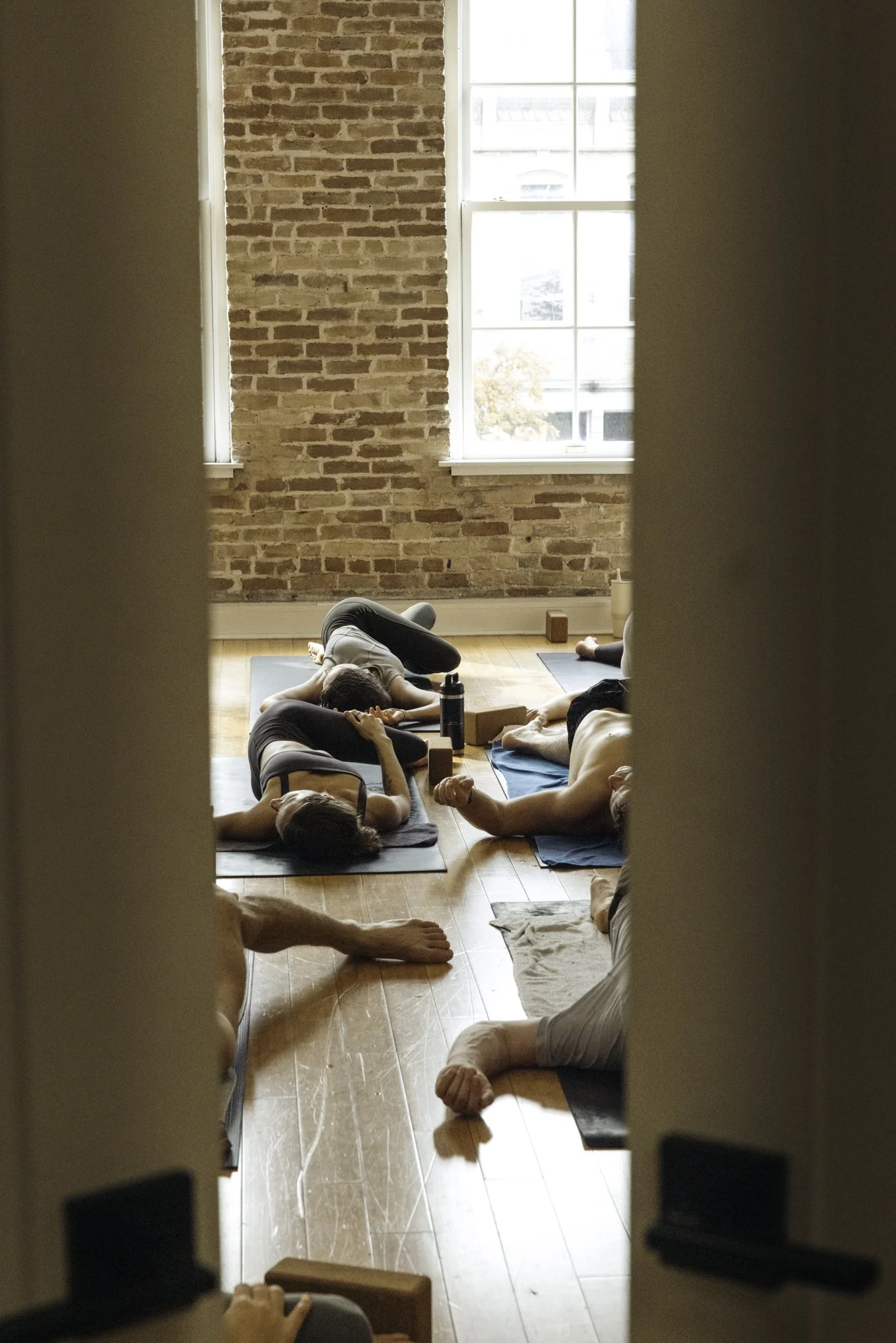 View through a slightly opened door into a cozy yoga studio where participants lie on their mats in a restful pose, surrounded by exposed brick walls and natural light.