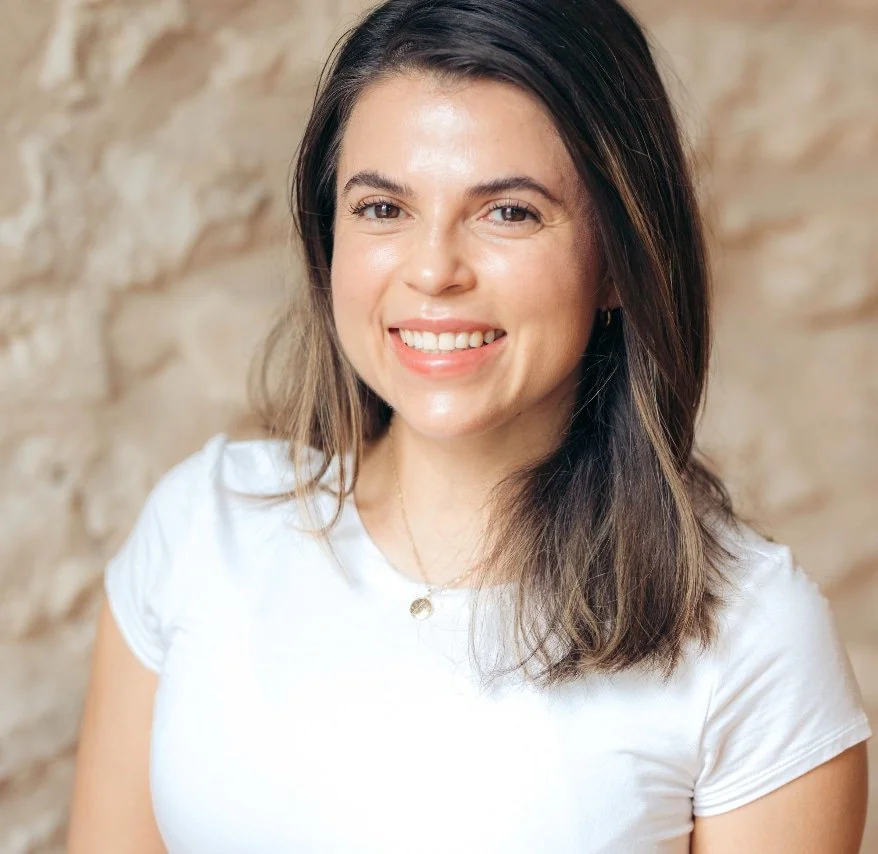 A smiling woman with shoulder-length brown hair wearing a white shirt and a delicate necklace against a beige stone wall.