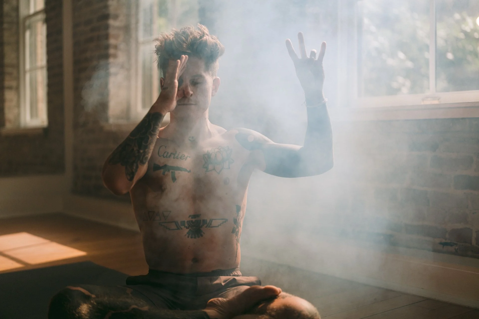 A shirtless young man with tattoos sitting on the floor in a yoga pose in a sunlit room with brick walls, surrounded by smoke or mist.
