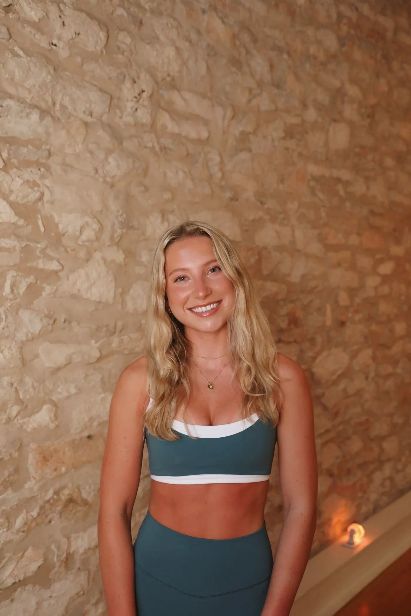A young woman with long blonde hair smiling, wearing a teal sports bra and matching leggings, standing in front of a beige stone wall.