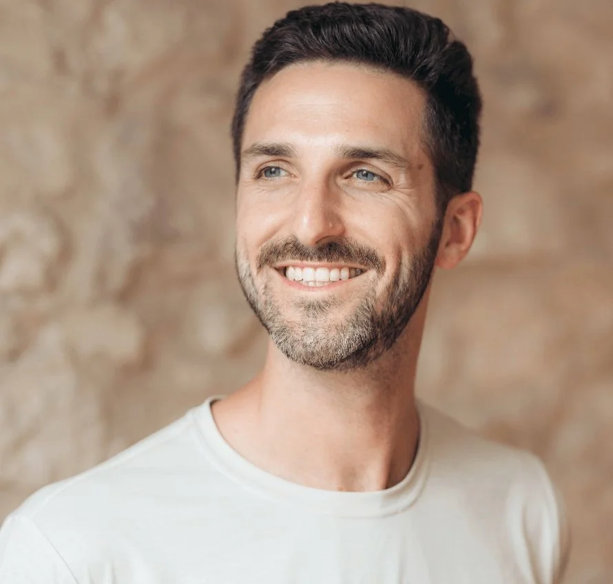 Close-up of a smiling man with short dark hair, facial hair, blue eyes, and wearing a white shirt, standing against a brick wall background.