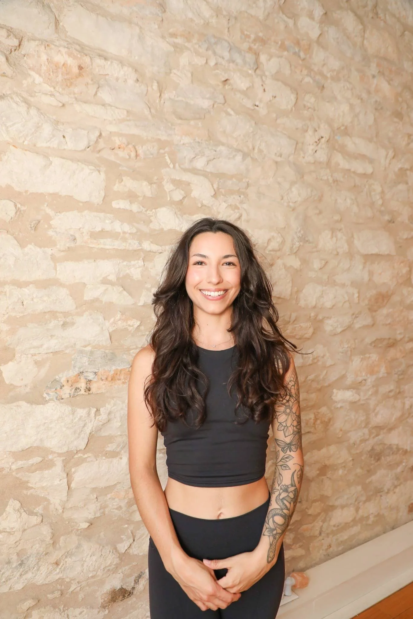 Woman with long wavy hair and a floral arm tattoo smiles warmly while standing against a textured stone wall, dressed in a black crop top and leggings.