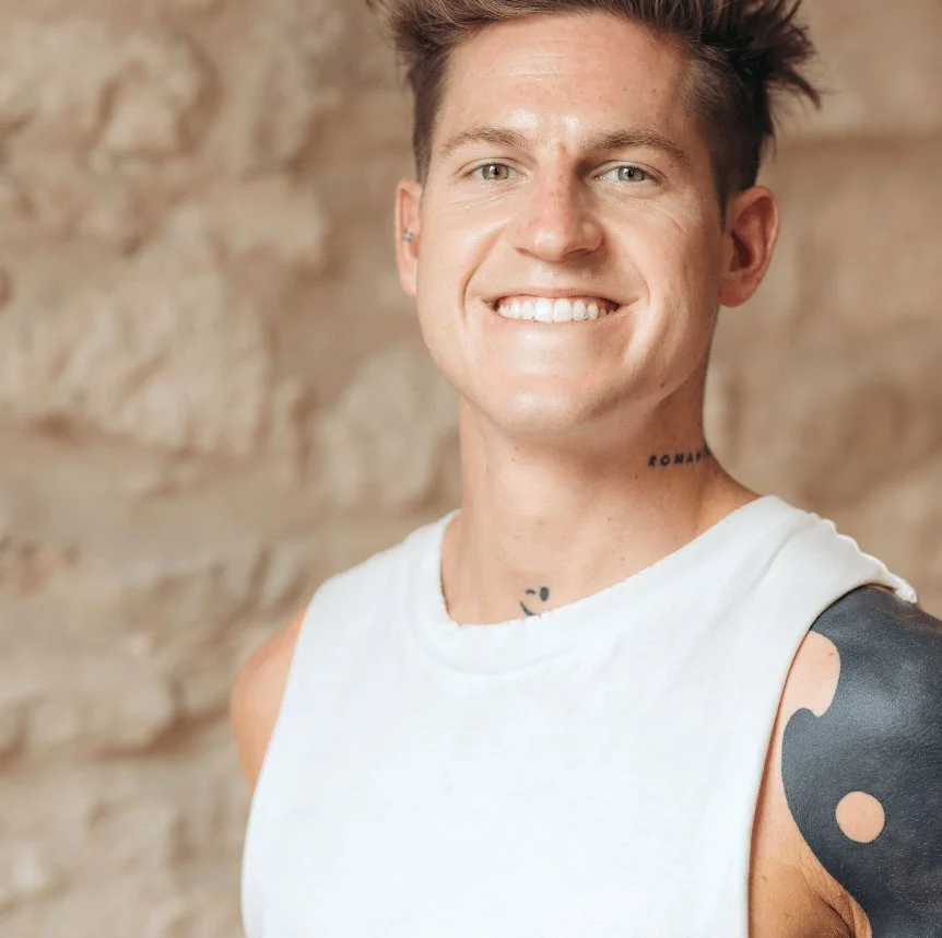 Smiling young man with short spiked hair wearing a white sleeveless shirt, standing in front of a textured beige wall, with tattoos on his neck and arm.