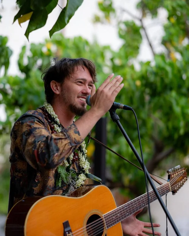 Young man with beard and lei holding a guitar, smiling into a microphone, with his hand reaching toward the microphone in a setting of leaves and greenery.