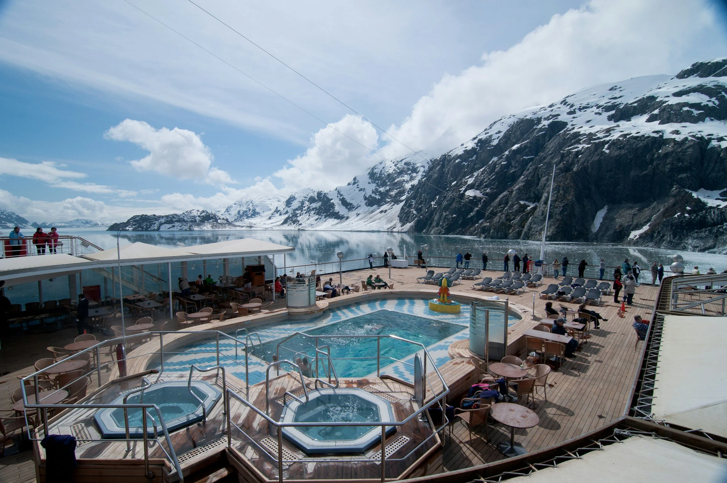 Cruise ship deck with hot tubs, lounge chairs, and a view of snow-covered mountains and water.