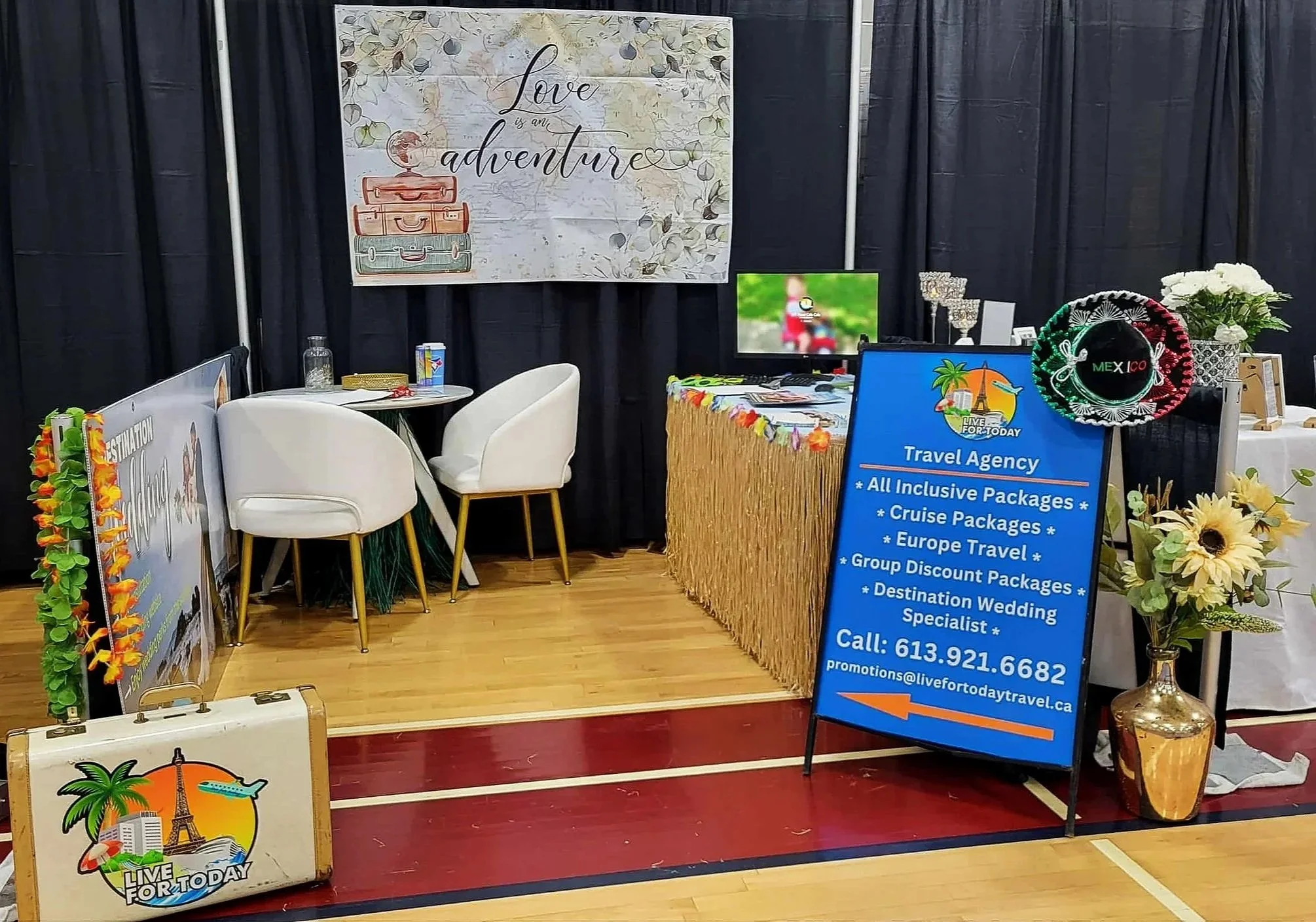 Travel agency booth with signs and promotional materials for vacation packages, decorated with tropical themes, sunflowers, and a large sign reading 'Love is an adventure'.
