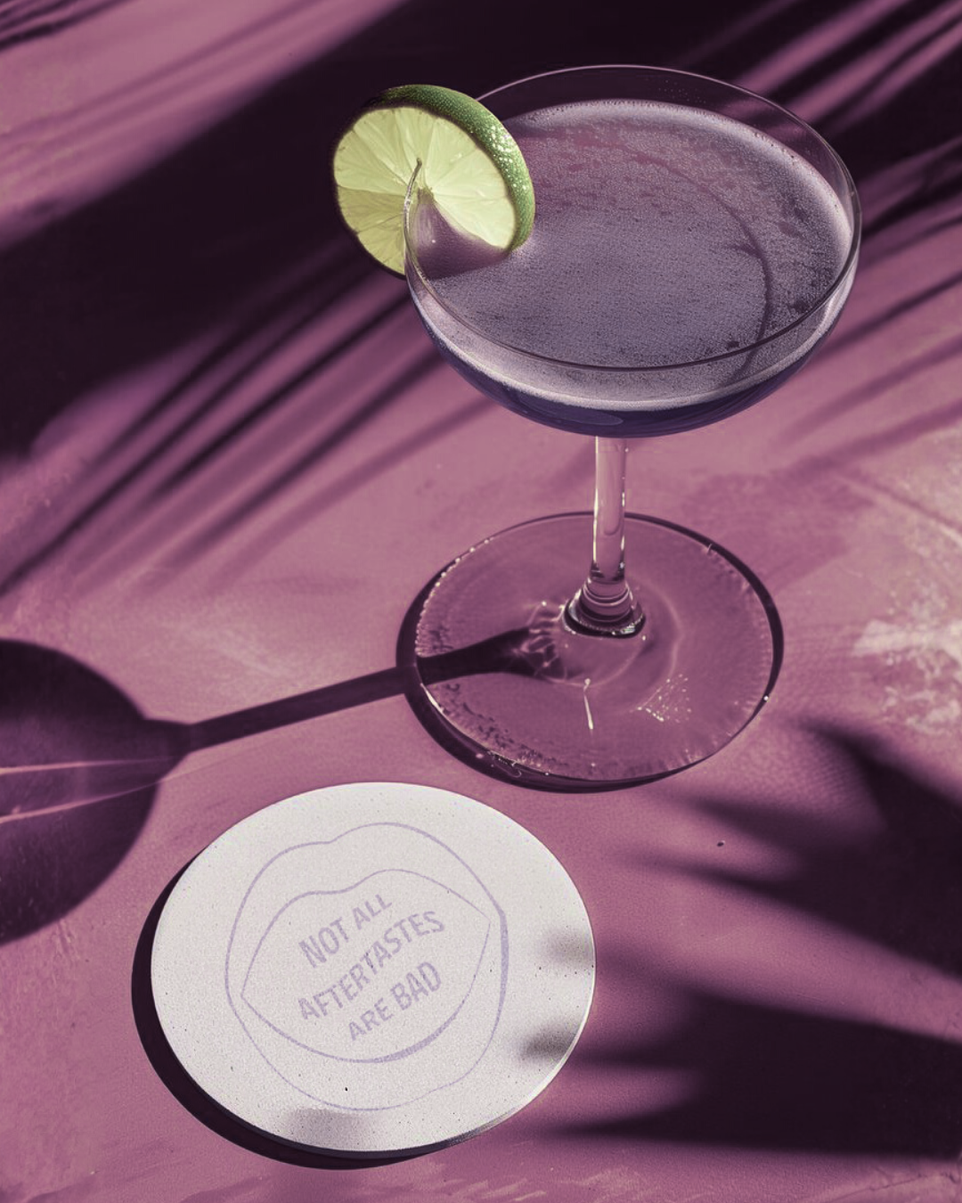 A cocktail glass with a lemon wedge slice on the rim, placed on a pink surface with shadows of leaves, and a round white coaster with the message 'Not all aftertastes are bad'.