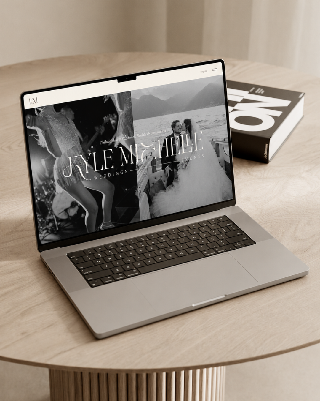 Open laptop displaying a wedding planning website with a black and white photo of a couple on a boat and a woman dancing at a party, placed on a round wooden table with a black and white book nearby.