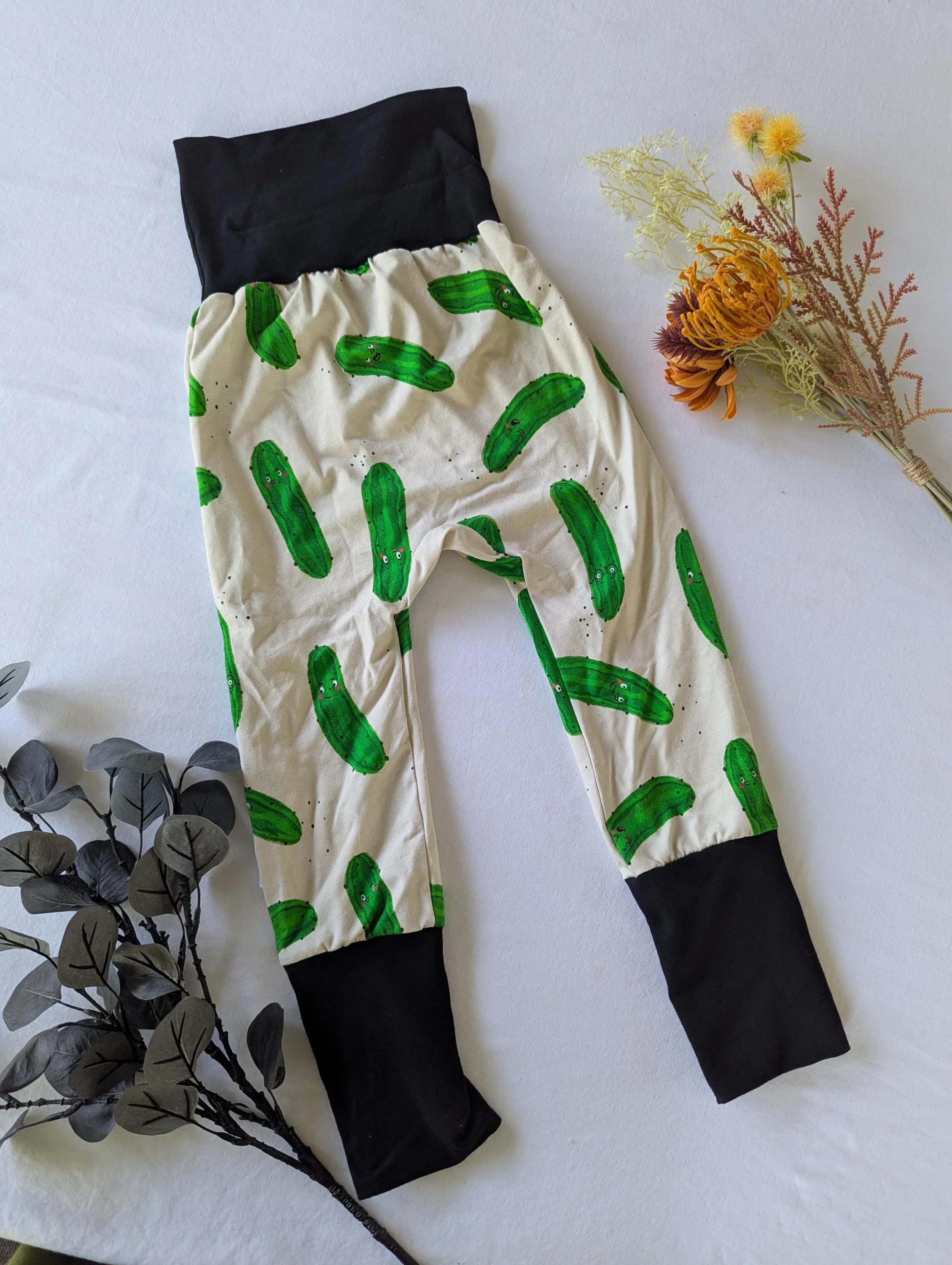 Pajamas with green cucumber print and black cuffs lying on a white surface, next to a dried flower arrangement and an artificial gray leaf branch.