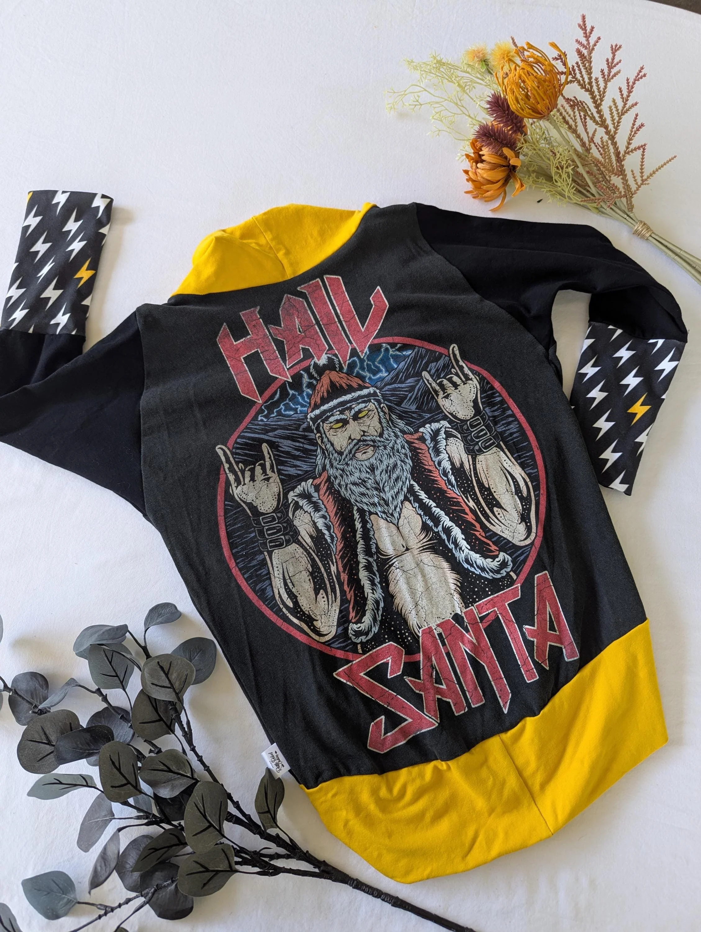 Black and yellow long-sleeve shirt with a graphic of a bearded, muscular man making rock hand signs, wearing a red cap and vest. The shirt is laid flat on a surface with a flower bouquet in the top right corner and a gray leafy branch in the bottom left corner.