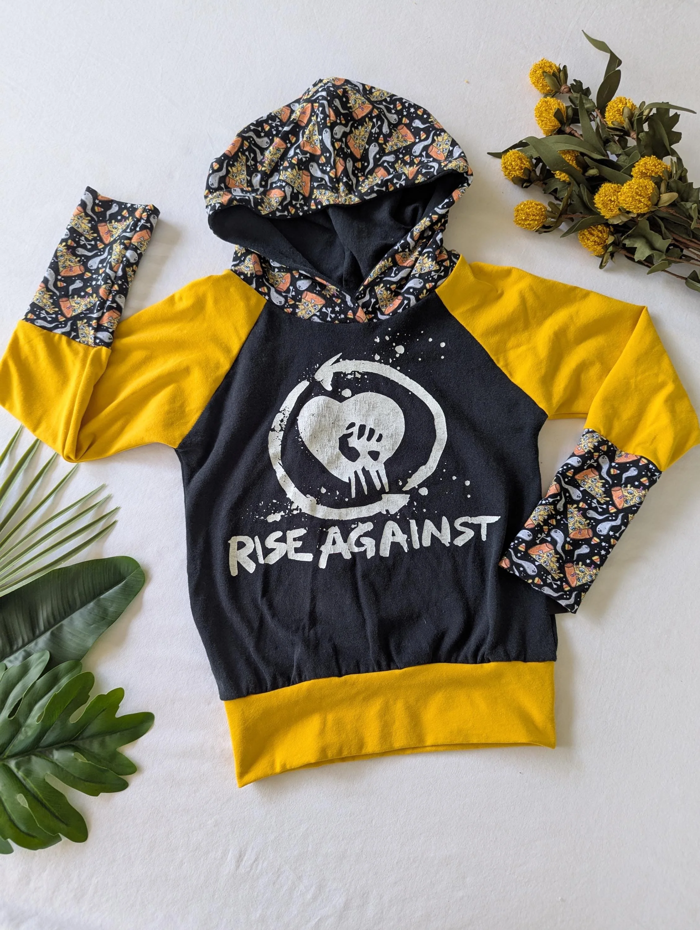 Black Children's hoodie with yellow sleeves- cuffs and hood have ghosts and pizza slices with eyeballs as toppings- white words 'rise against' with band's heart logo on torso front
