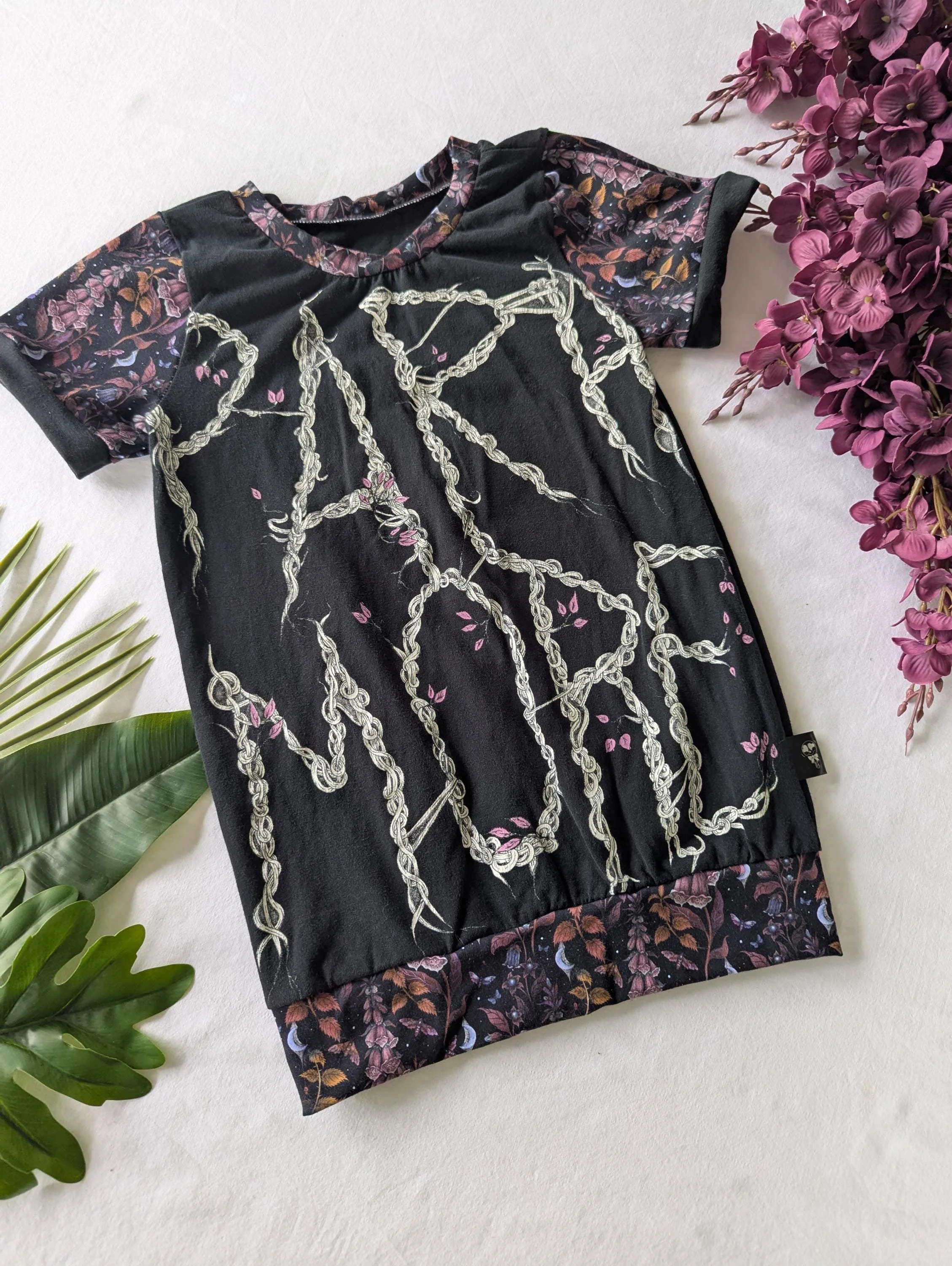 A black t-shirt with a floral and chain design, surrounded by green and purple leaves and purple flowers.