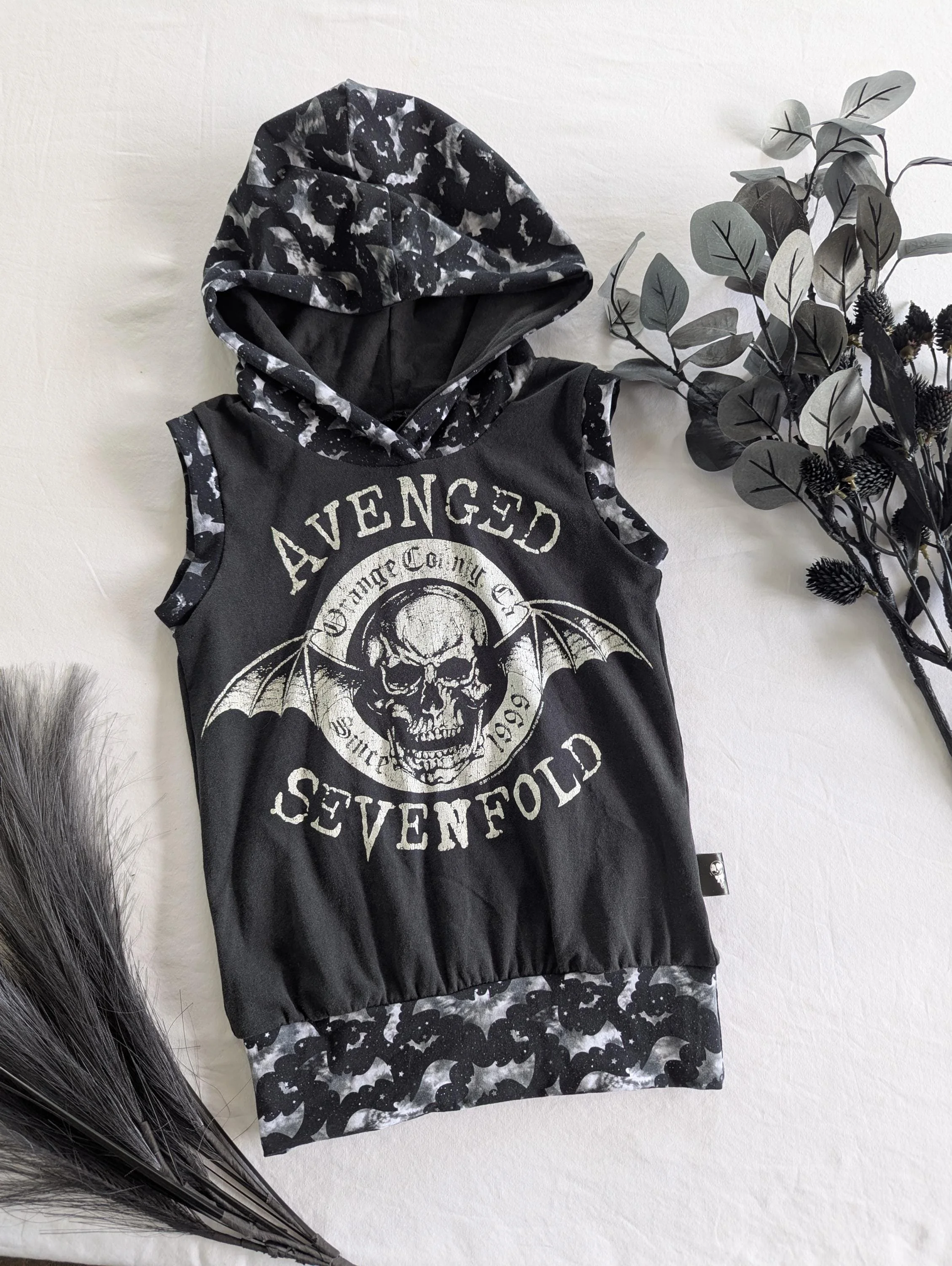 A sleeveless black hoodie with a skull and bat wings graphic and the words "Avenged Sevenfold" and "Since 1999" printed on the front, with a matching camo hood.
