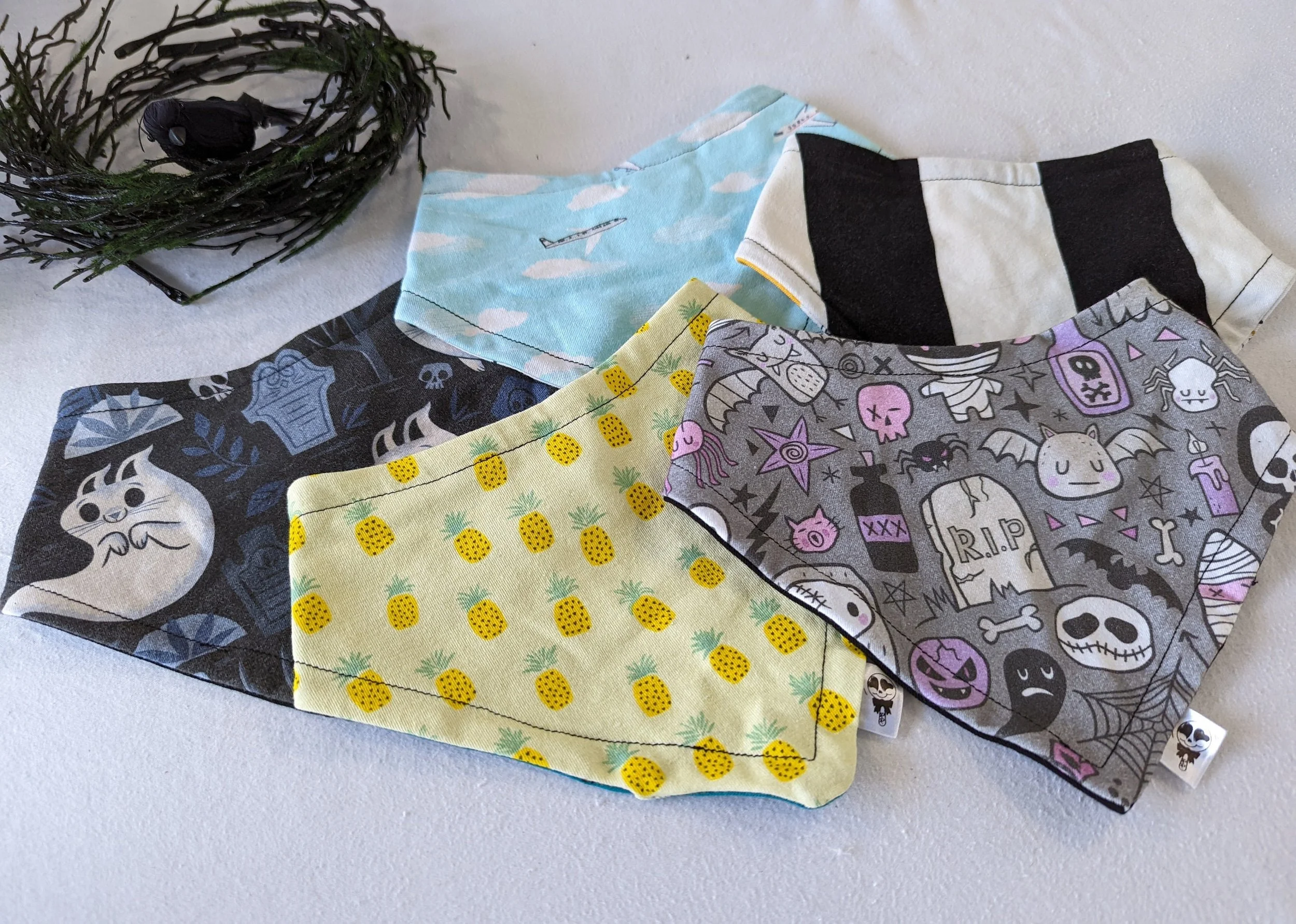 Five fabric bibs with different patterns, spread on a white surface, with a decorative wreath in the background.
