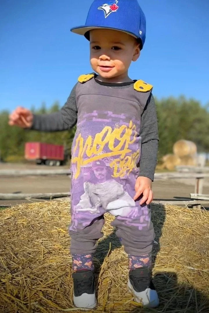 A young boy wearing a Toronto Blue Jays cap, a gray shirt with yellow shoulder patches, and purple pants featuring a graphic of a woman in a yoga pose, standing on a hay bale outdoors on a clear day.