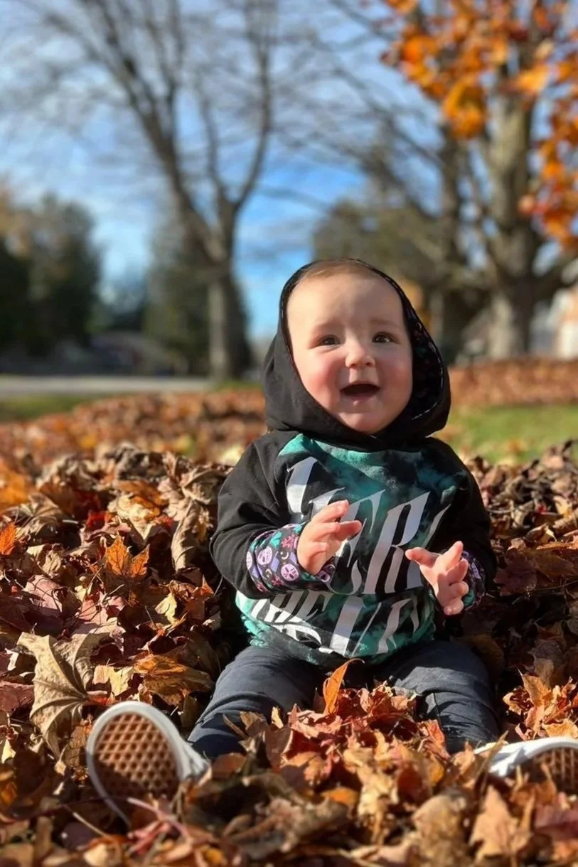 A Baby Sitting in a pile of autumn leaves outdoors with trees in the background and a blue sky.