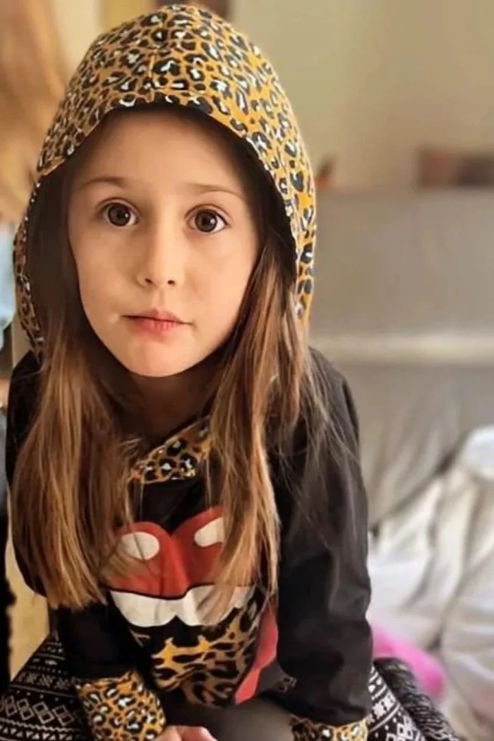 A young girl with long brown hair and big brown eyes, wearing a leopard print hoodie and a black sweatshirt with a red tongue logo, sitting indoors.
