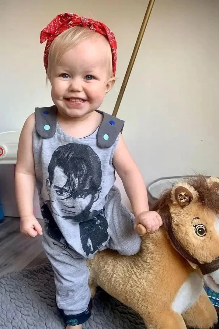 Happy toddler girl wearing a red bandana on her head, sitting on a plush toy horse, smiling with teeth showing, indoors with a plain background and a power outlet in the corner.