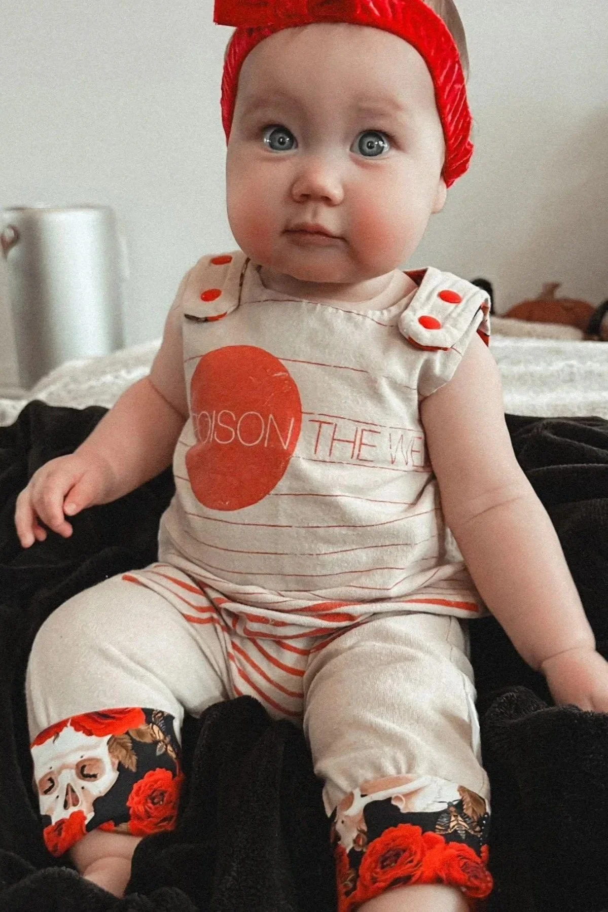 Young child wearing a red headband and a cream-colored outfit with skull and floral designs, sitting on a black blanket.