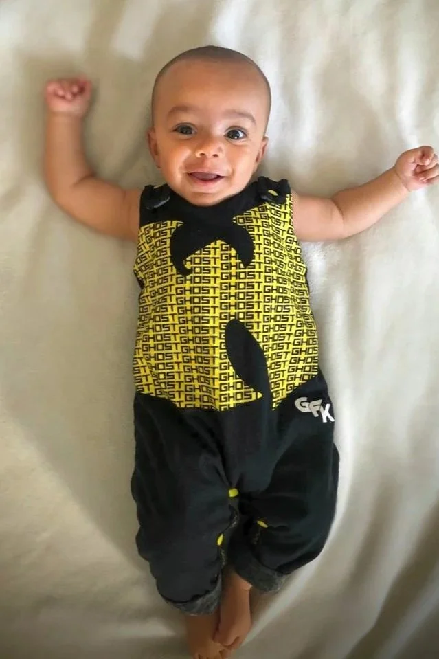 Smiling baby lying on a bed with arms outstretched, wearing a yellow and black Ghostbusters-themed outfit.
