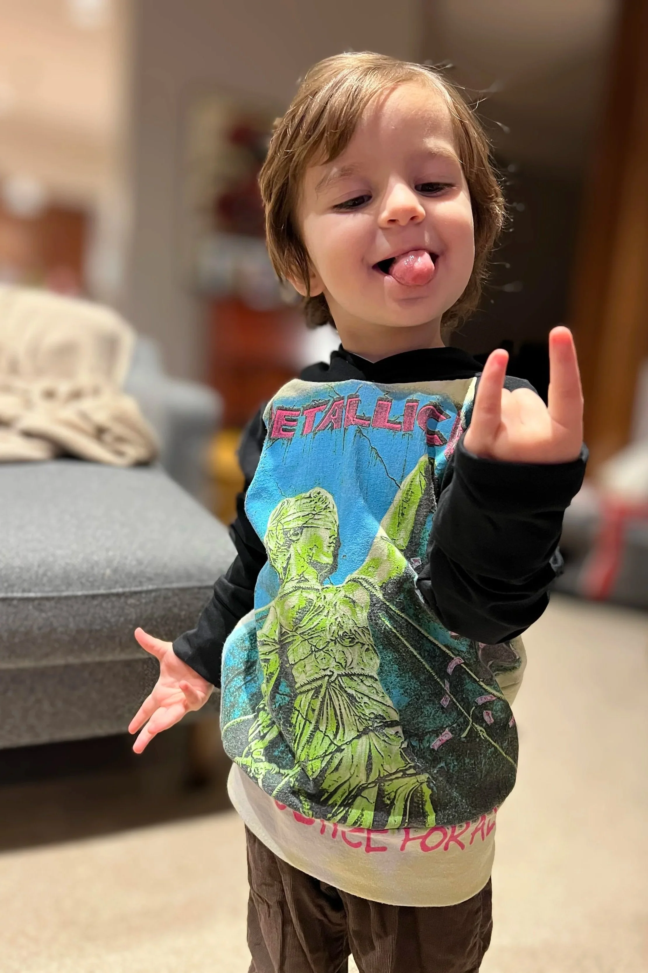 A young boy with light brown hair sticking out his tongue and showing his middle finger, wearing a Metallica band t-shirt, standing indoors in front of a blurred background.