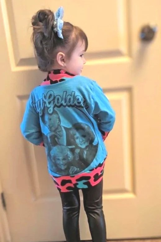 Little girl with a large blue jacket featuring vintage photos of women and the word 'Goldie' on the back, wearing black shiny pants and a blue hair bow, standing in front of a beige door.