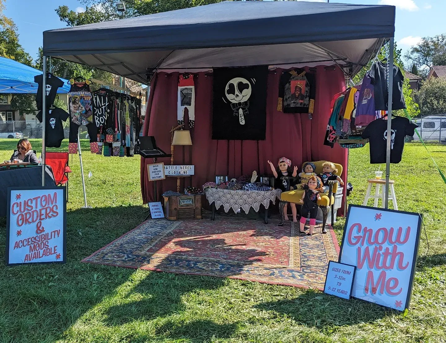 A street vendor booth selling custom grip clothing and children's clothing at an outdoor event. Signs indicate customization options and sizes for children aged 3-12 years. The booth has a vintage style rug, a mannequin, toys, and colorful clothing displayed.