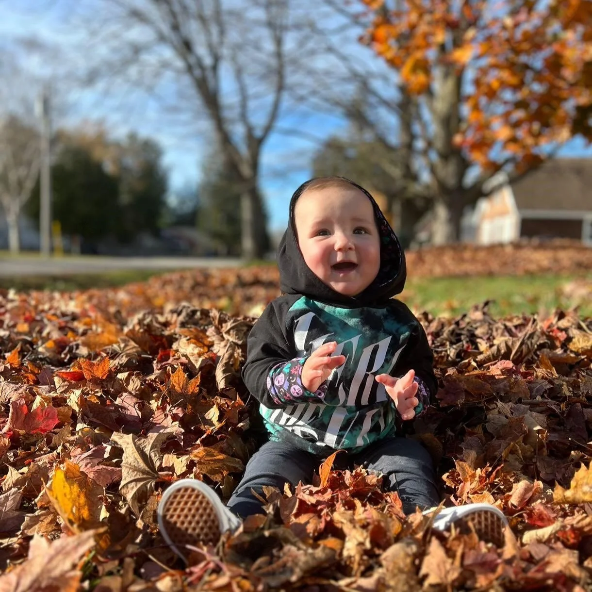 A young child sitting in a pile of autumn leaves outdoors on a sunny day, smiling and clapping. There are trees with autumn foliage and houses in the background.