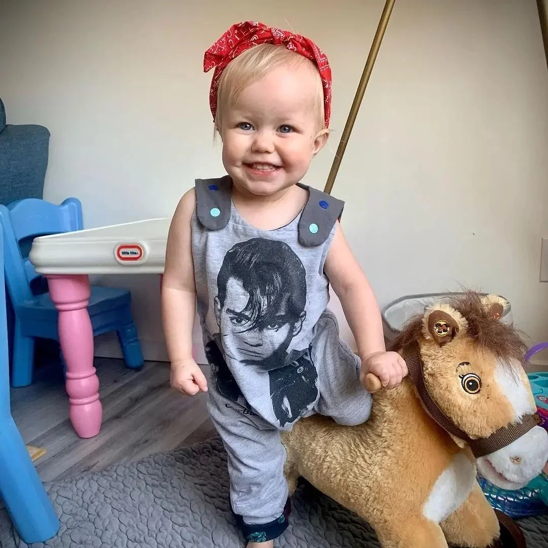 A smiling toddler wearing a gray tank top with a graphic of a man with sunglasses, gray pants, a red bandana headscarf, and small blue shoulder details, riding a plush toy horse in a room with a blue and white child's table and chair, and a beige wall.