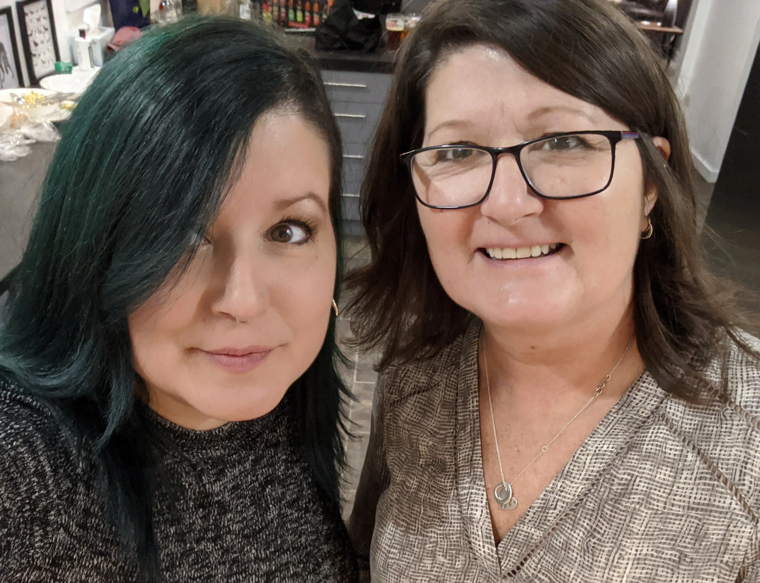 Two women smiling for a selfie in a kitchen, one with dark hair and glasses, the other with black hair and a nose piercing.